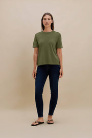 A woman modeling a short-sleeve crewneck t-shirt in olive green, paired with dark blue skinny jeans and black flip-flops, standing against a solid beige studio background.