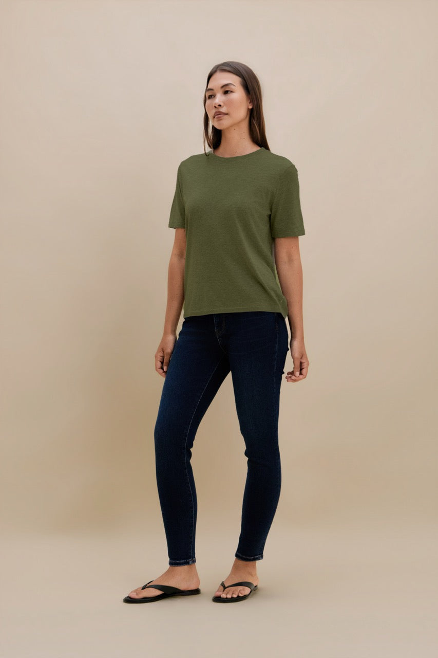 A woman modeling a short-sleeve crewneck t-shirt in olive green paired with dark blue skinny jeans and black flip-flops, standing against a solid beige studio background.