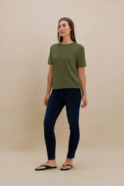 A woman modeling a short-sleeve crewneck t-shirt in olive green paired with dark blue skinny jeans and black flip-flops, standing against a solid beige studio background.