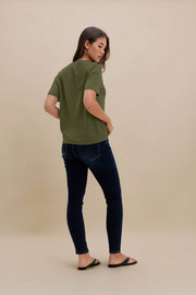 A woman wearing a short-sleeve olive green t-shirt and dark blue skinny jeans, photographed from the back against a solid beige studio background.