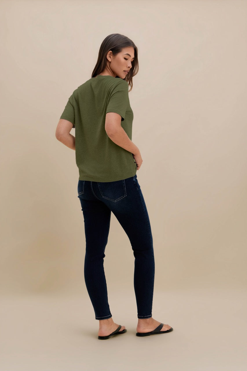A woman wearing a short-sleeve olive green t-shirt and dark blue skinny jeans, photographed from the back against a solid beige studio background.