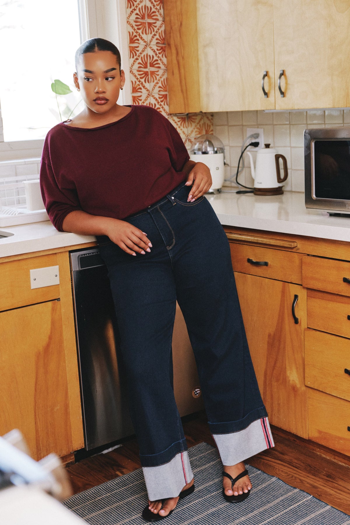 A woman stands in a kitchen wearing dark wide-leg jeans with rolled cuffs showing red  stripe detailing, paired with a burgundy top and black sandals.
