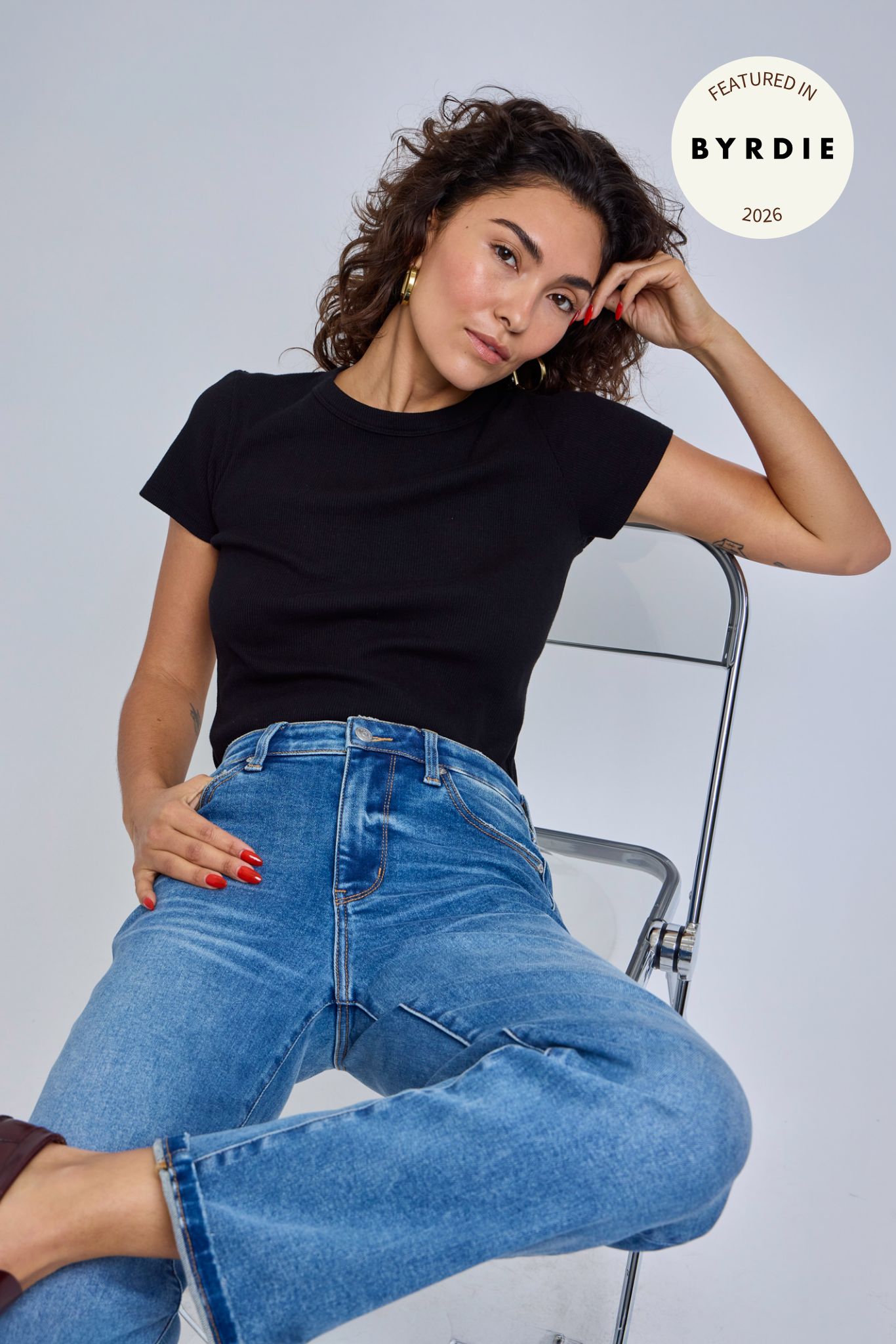 Woman wearing a black top and blue jeans sitting on a chair against a plain background | model-size: 4
