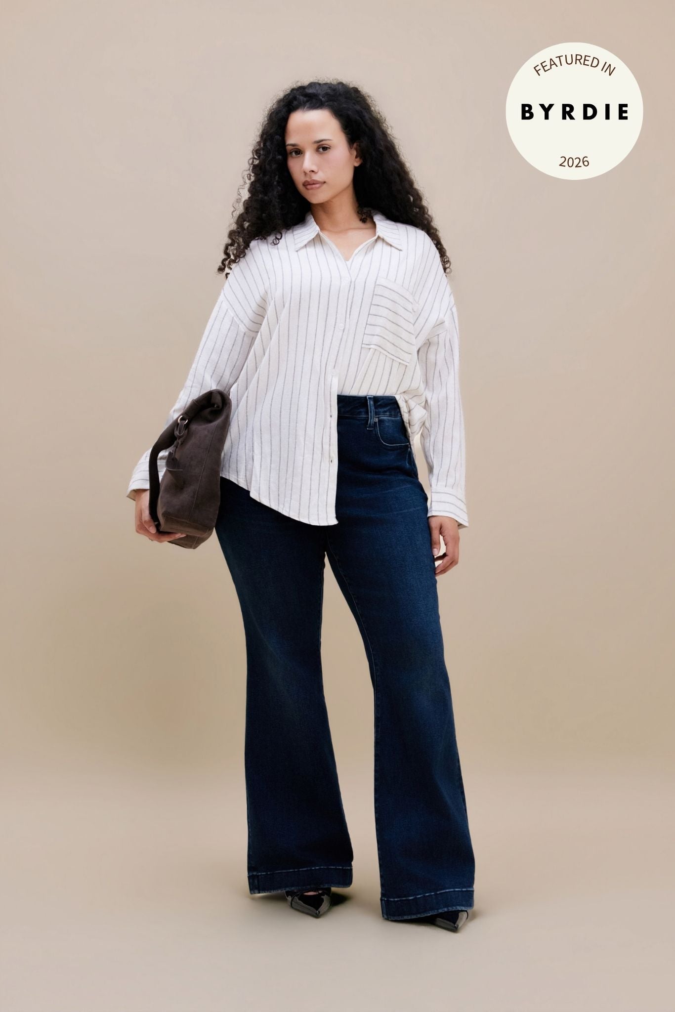 Woman wearing a white striped button down and dark flare jeans with a beige background and BYRDIE logo | model-size: 16