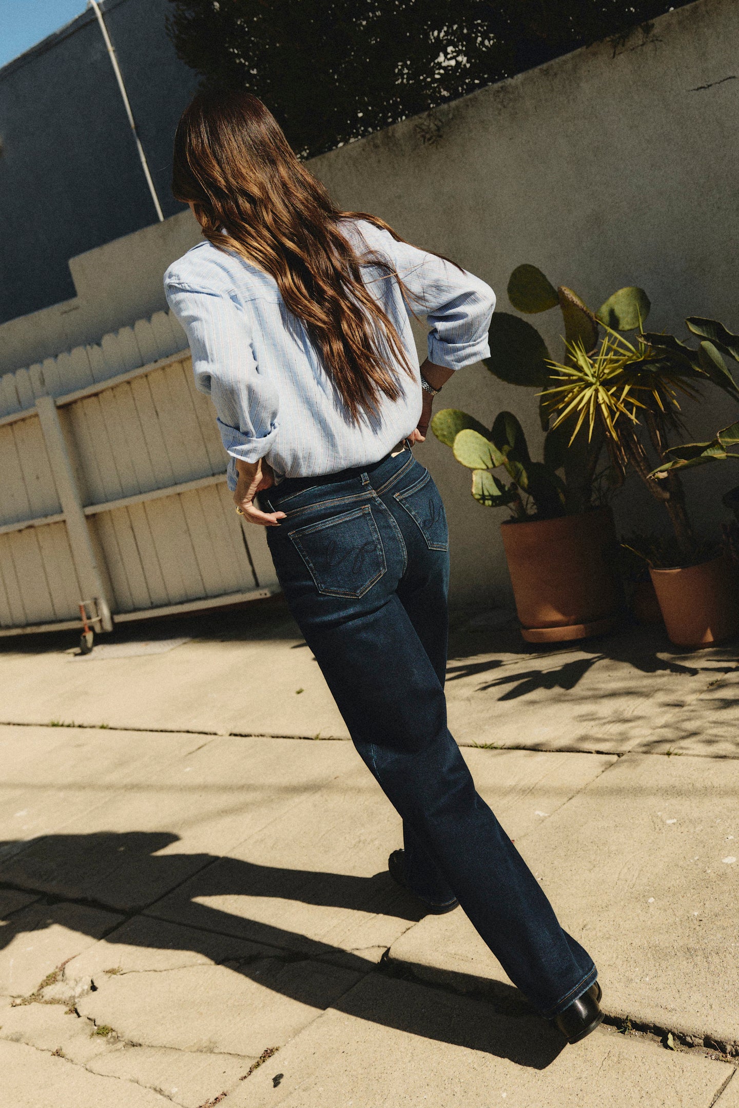 Person wearing a light blue shirt and dark jeans standing on a concrete surface with plants in the background | model-size: 4
