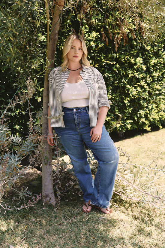 Woman standing outdoors in a garden wearing a striped shirt and blue jeans | model-size: 16