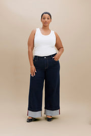 A woman modeling a pair of dark wide-leg denim pants with rolled cuffs, paired with a white tank top and black flats, against a solid beige studio background.