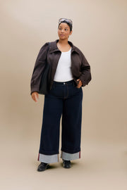 A woman modeling a dark brown cropped jacket, dark wide-leg jeans with rolled cuffs, a white tank top, and black shoes against a solid beige studio background.