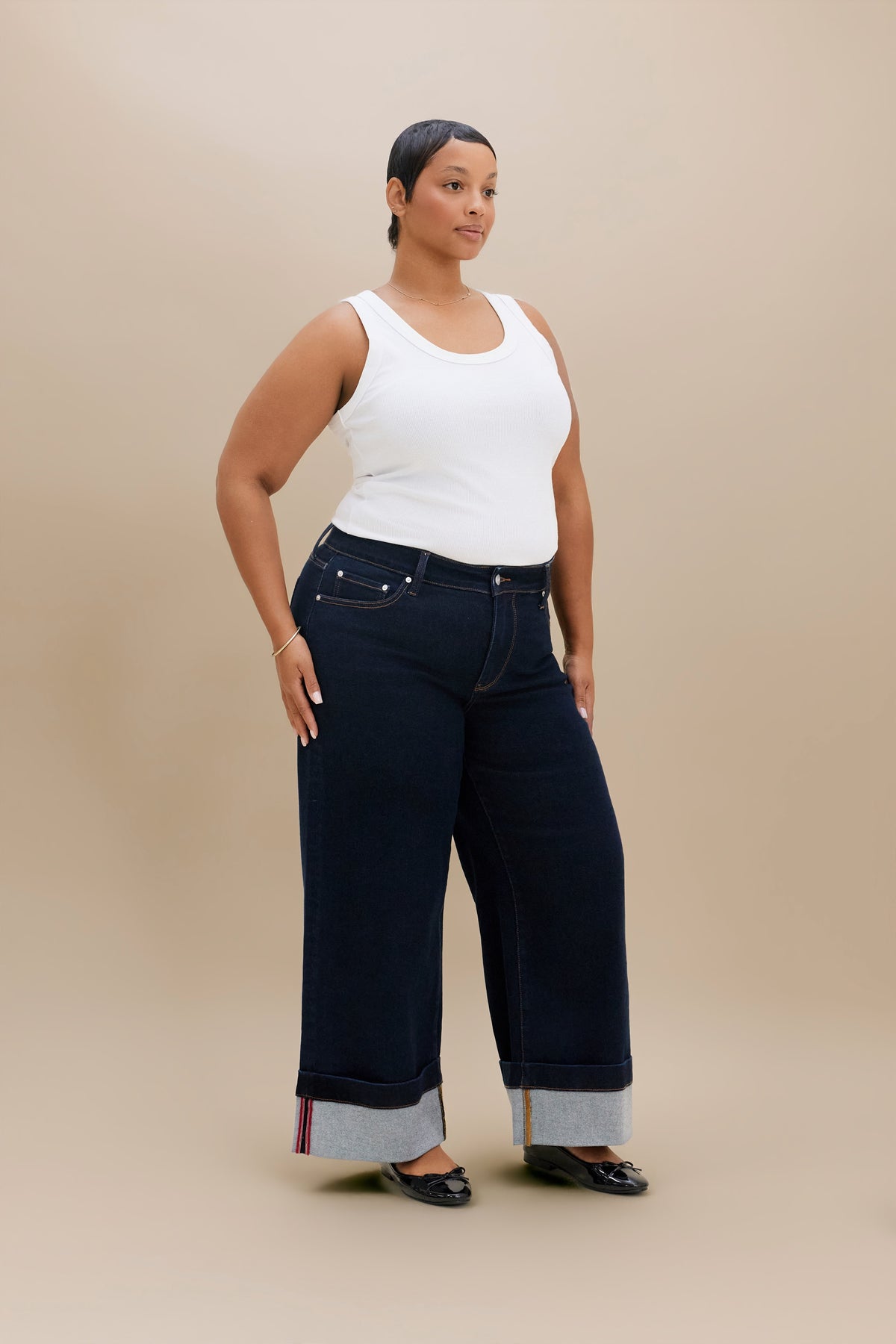 A woman modeling dark wide-leg jeans with rolled cuffs showing a light inner lining, paired with a white tank top and black flats, against a solid beige studio background.