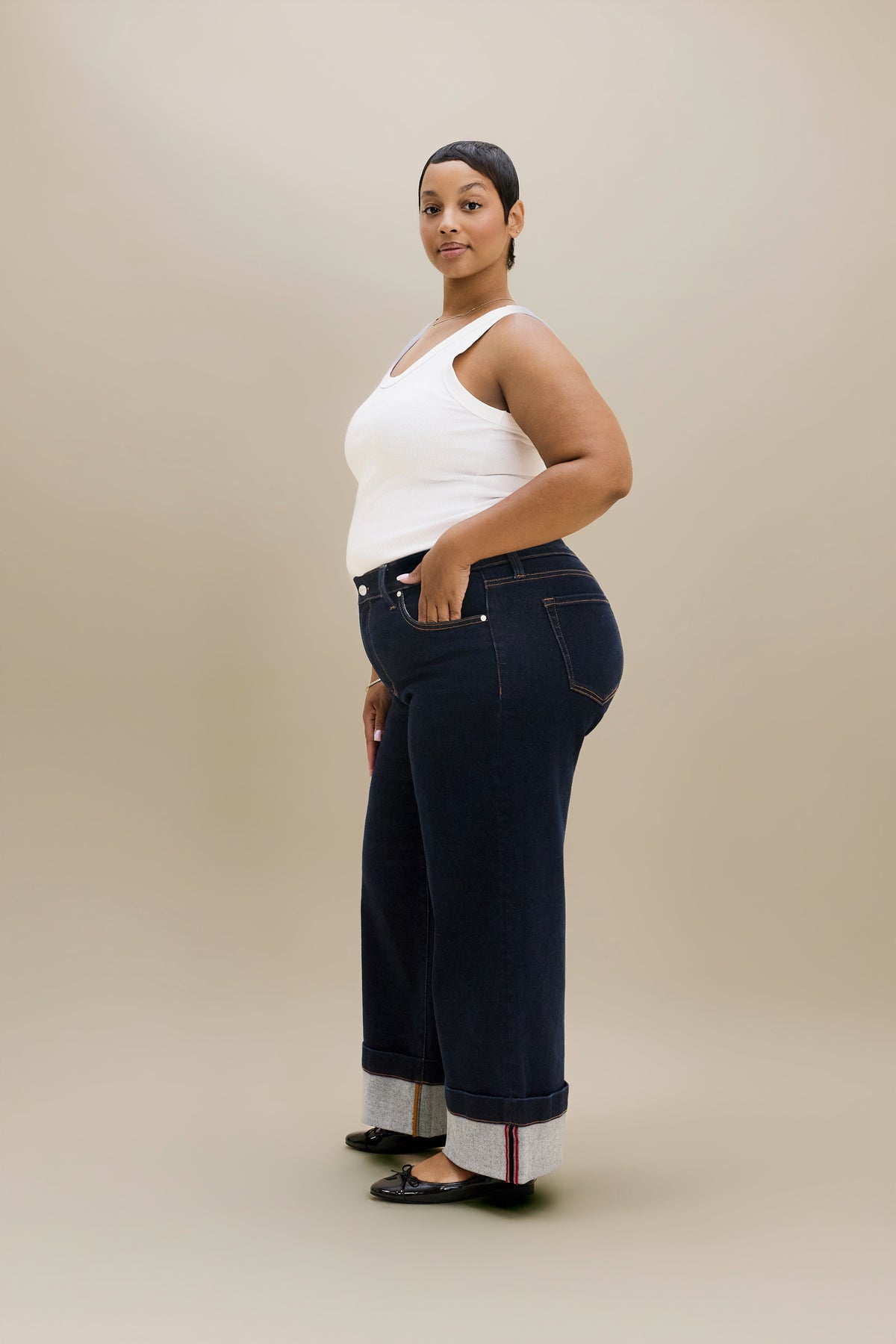 A woman modeling dark blue wide-leg jeans with rolled cuffs showing a white inner lining, paired with a white tank top and black flats, standing against a solid beige studio background.