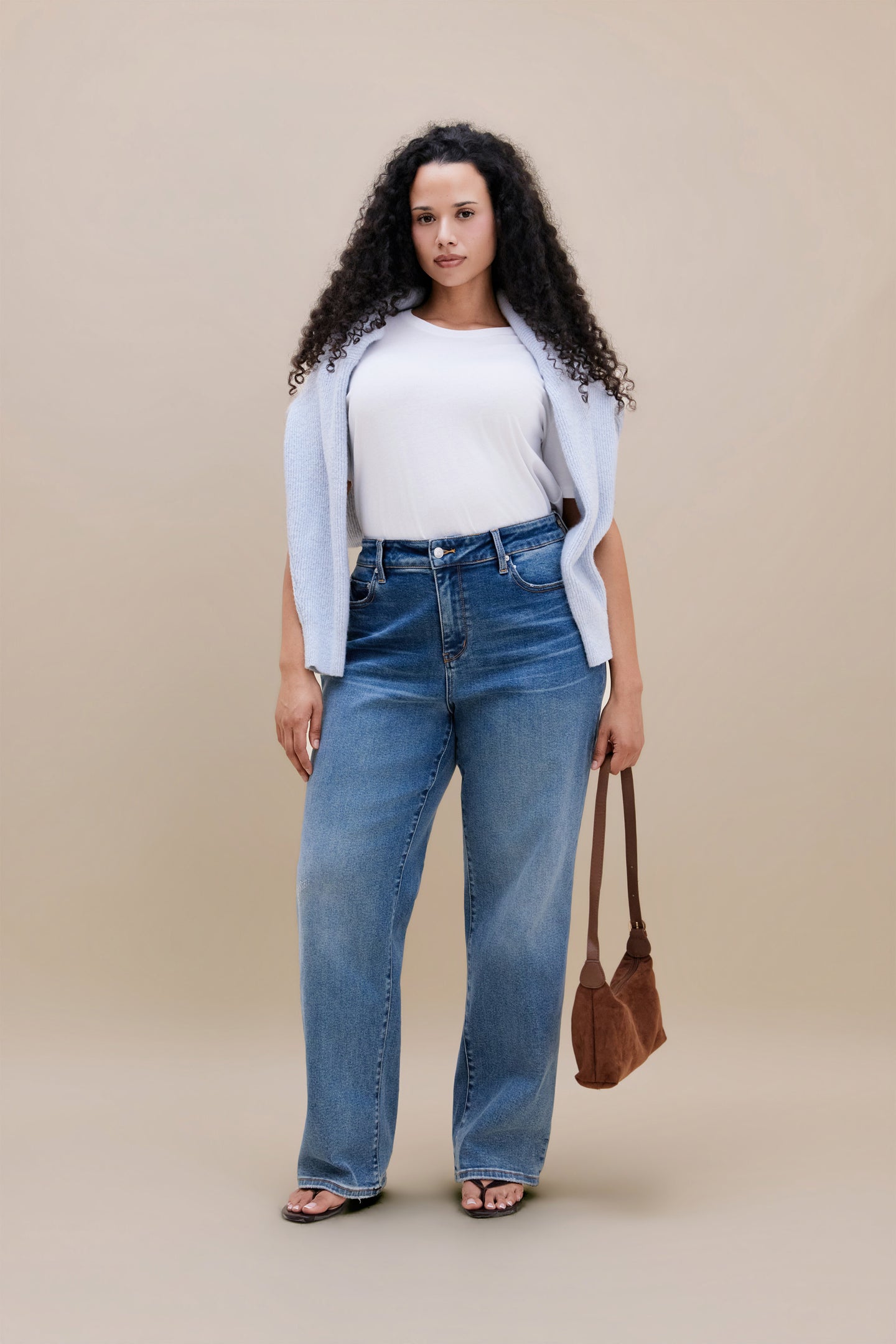 Woman wearing white t-shirt, blue sweater wrapped around shoulder and jeans holding a purse with beige background | model-size: 16