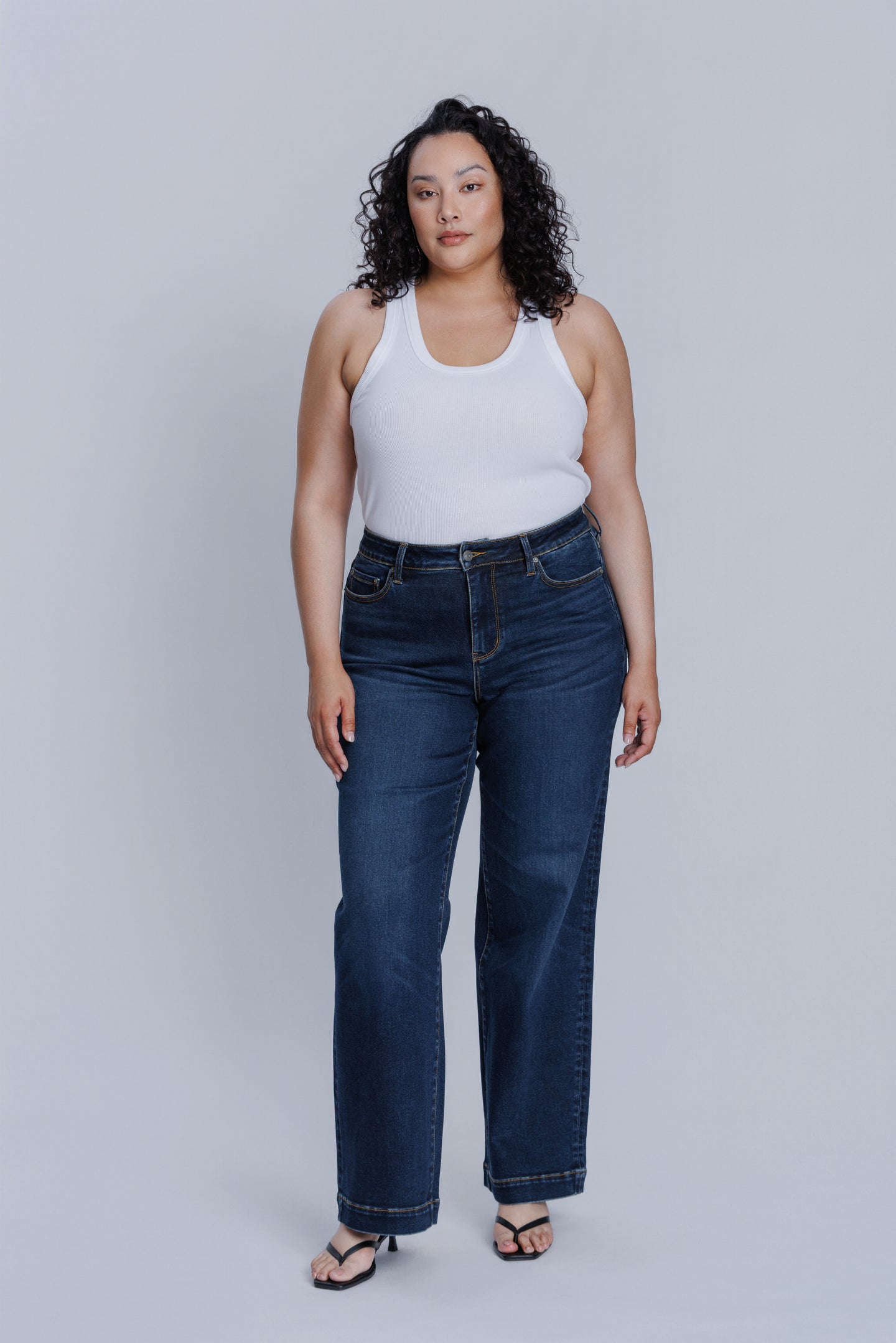 Woman wearing a DePart white tank top and dark blue jeans against a light gray background | model-size: 16