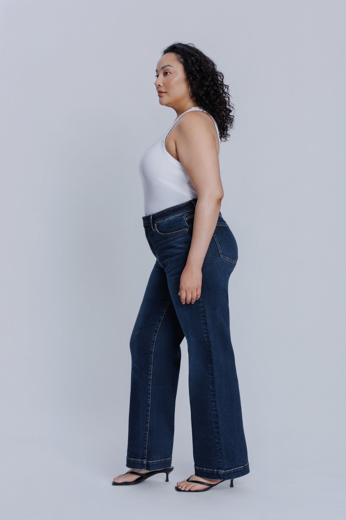 Woman wearing a DePart white tank top and dark blue jeans against a light gray background