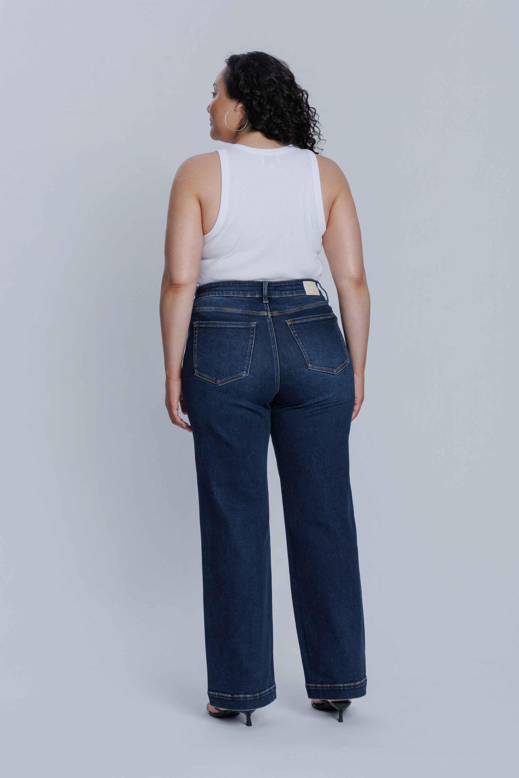 Woman wearing a DePart white tank top and dark blue jeans against a light gray background