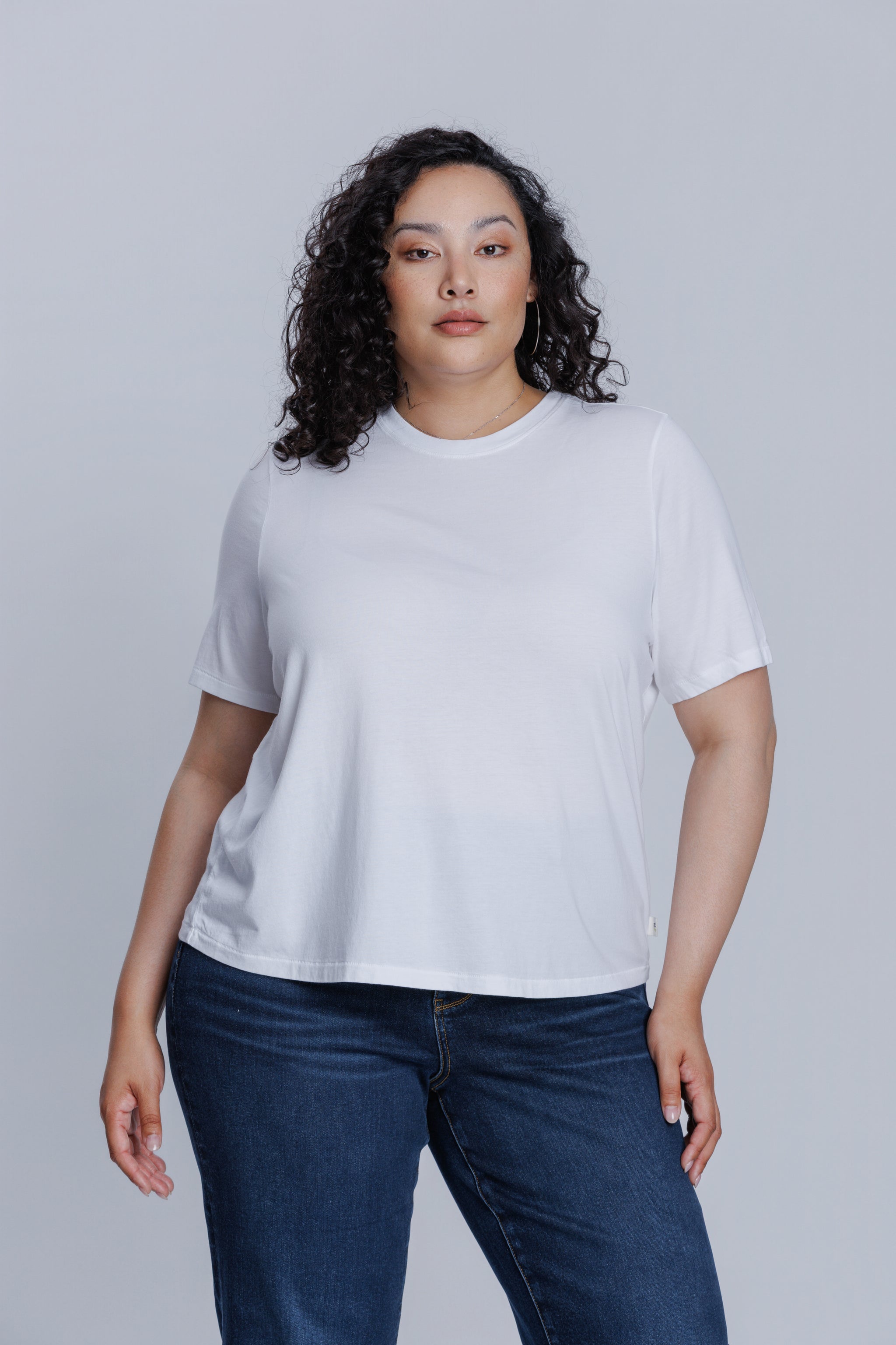Woman wearing a DePart white t-shirt and dark blue jeans against a light gray background | model-size: 16