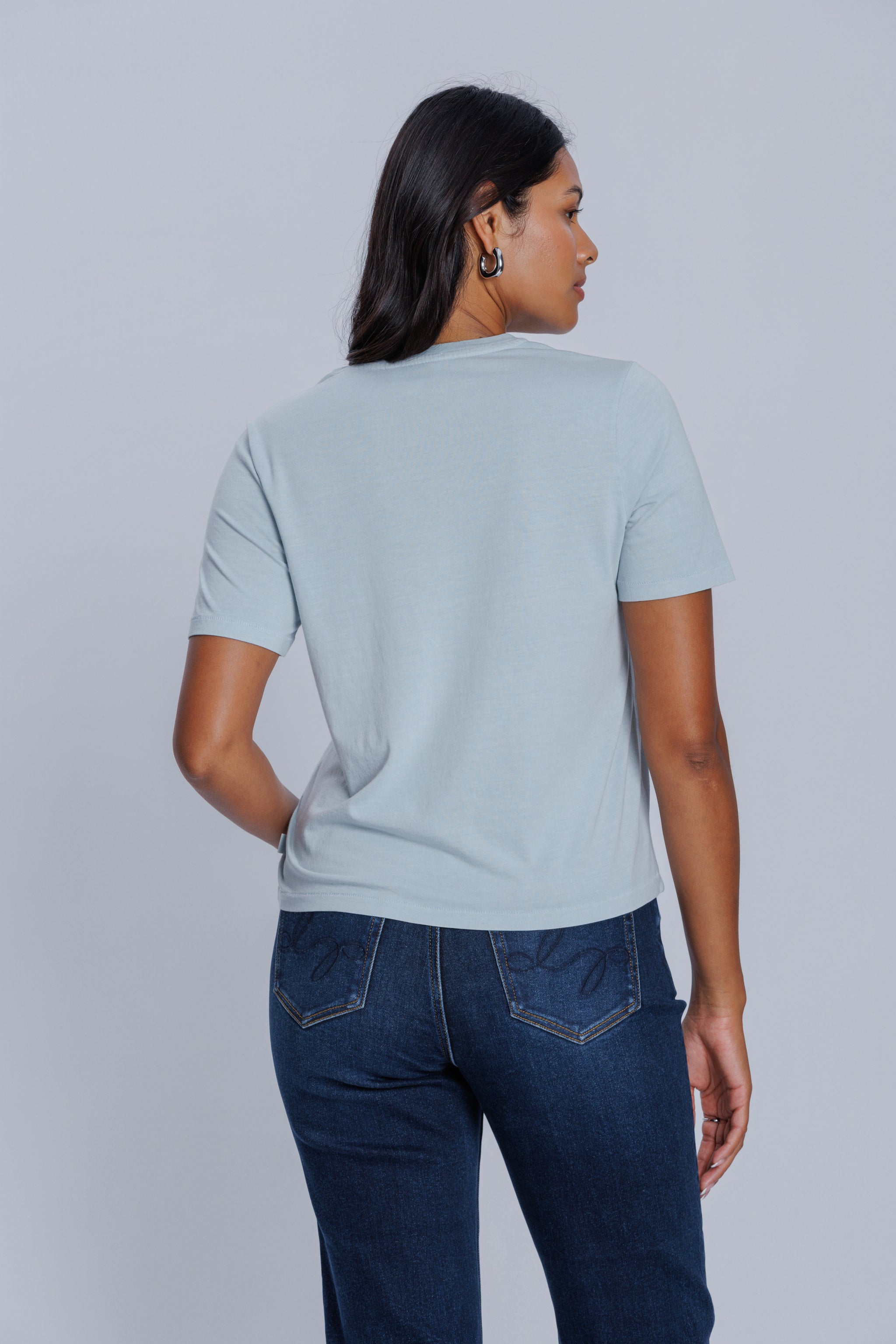 Woman wearing a DePart light blue t-shirt and dark blue jeans against a light gray background
