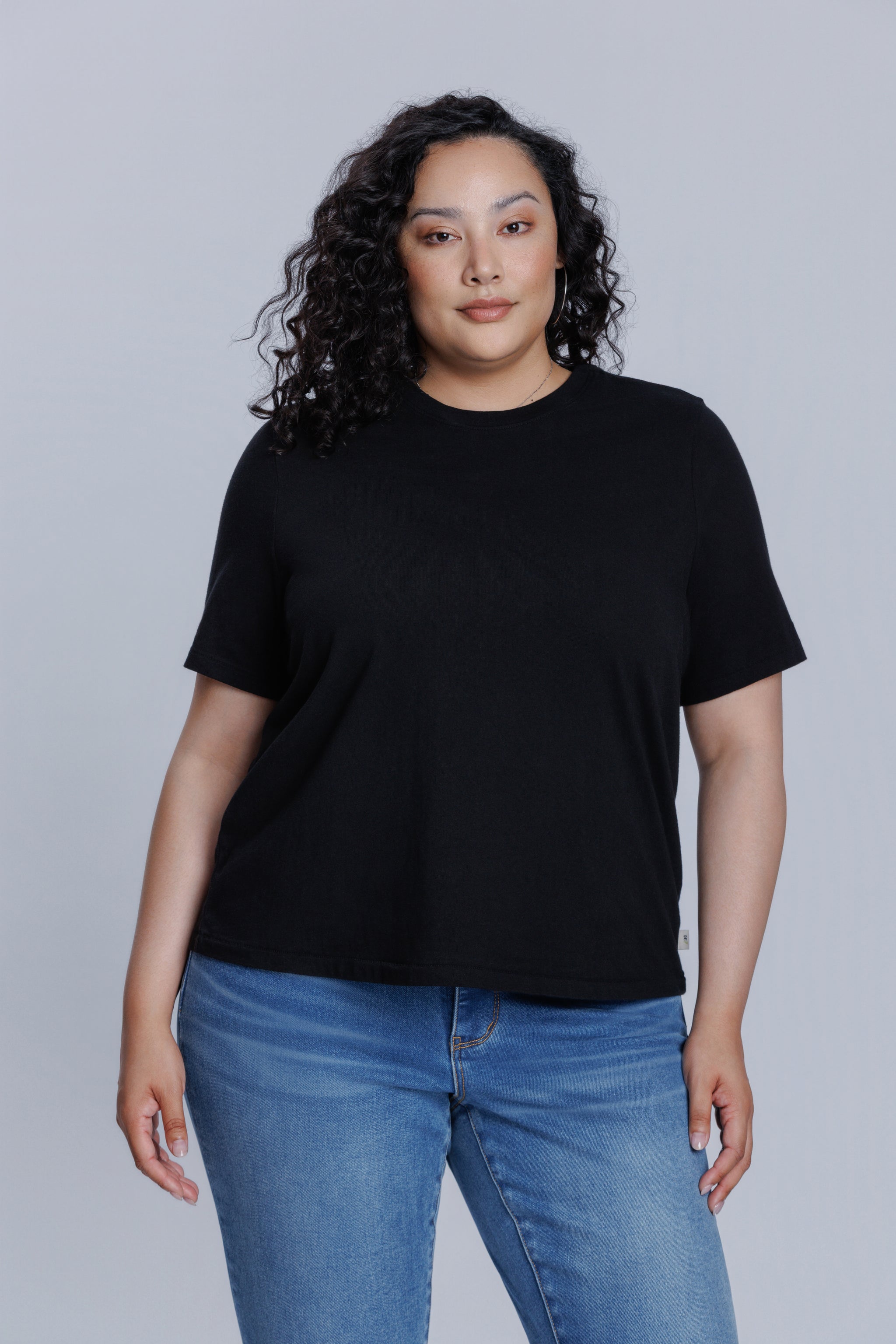 Woman wearing a DePart black t-shirt and blue jeans against a light gray background | model-size: 16