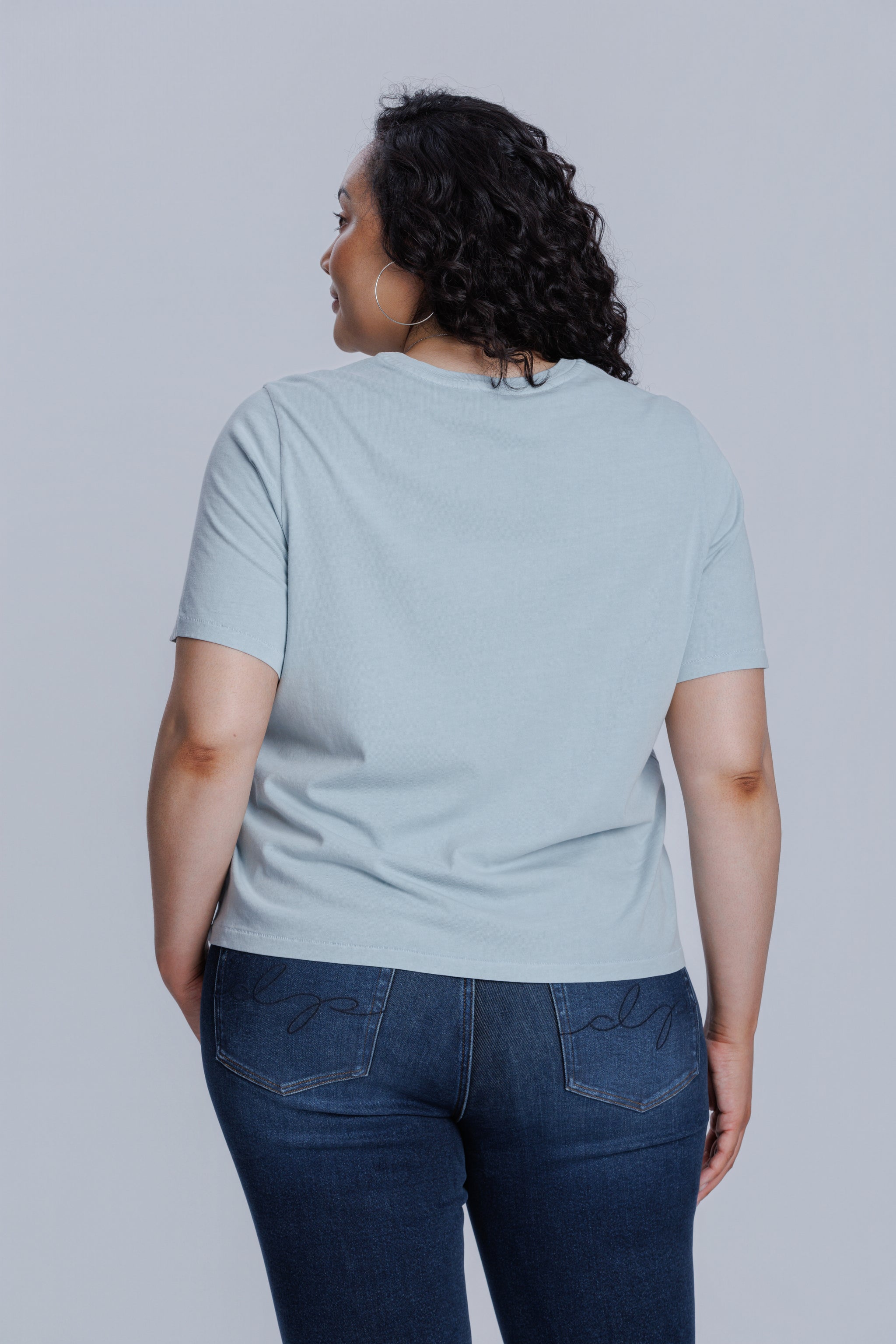 Woman wearing a DePart light blue t-shirt and dark blue jeans against a light gray background