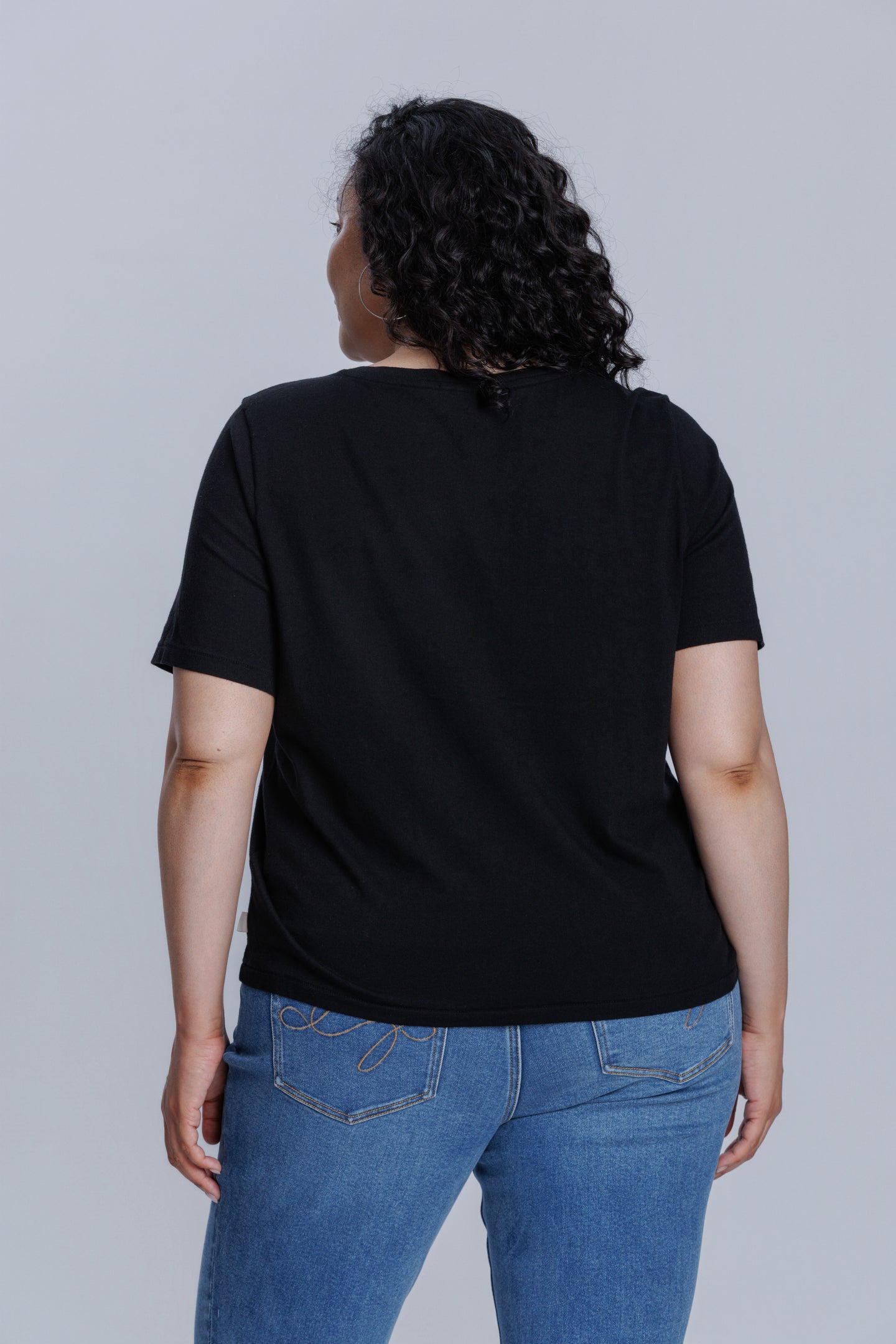 Woman wearing a DePart black t-shirt and blue straight jeans against a light gray background
