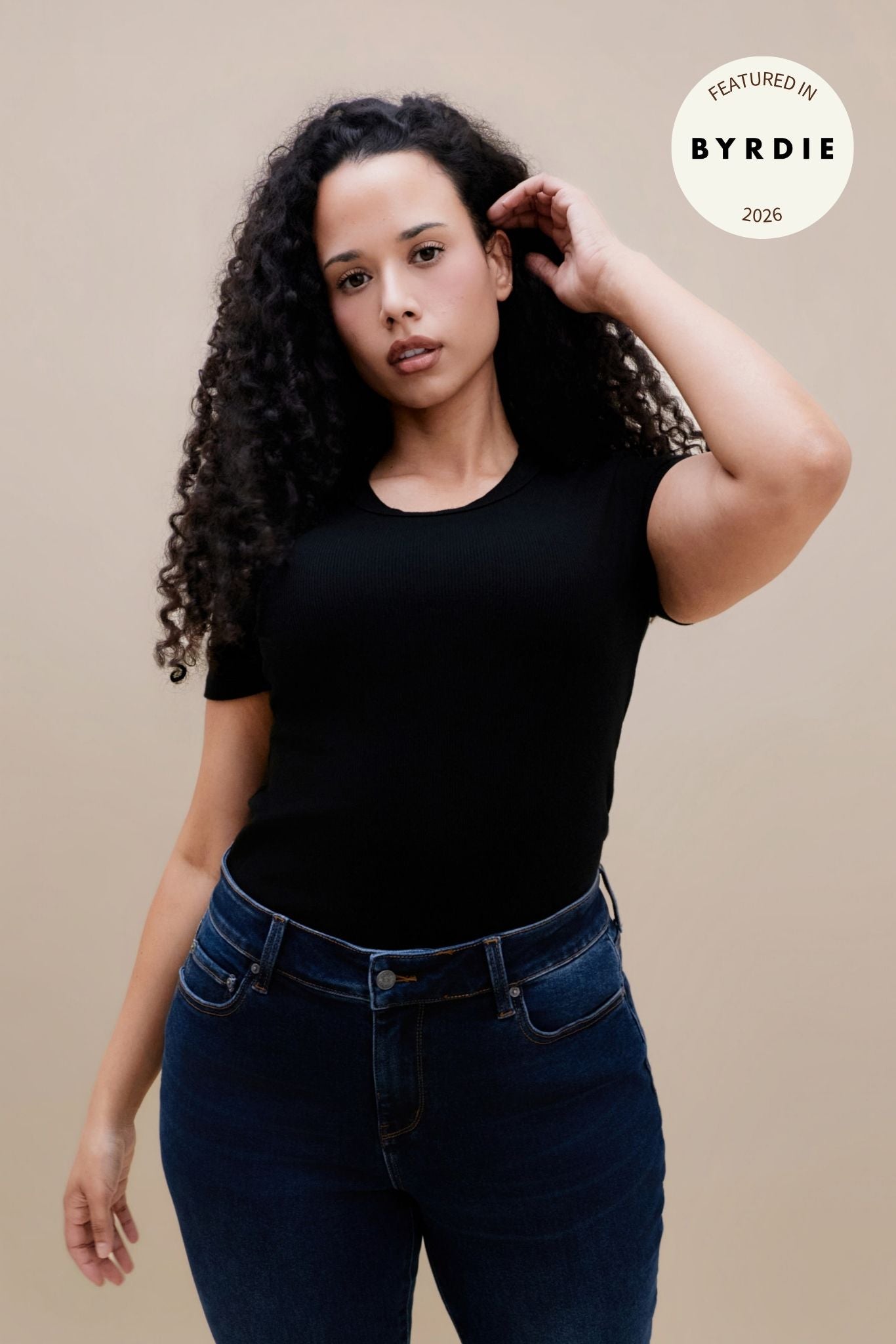 Woman wearing a fitted black rib tee and dark jeans against a beige wall with a 'BYRDIE' logo | model-size: 16