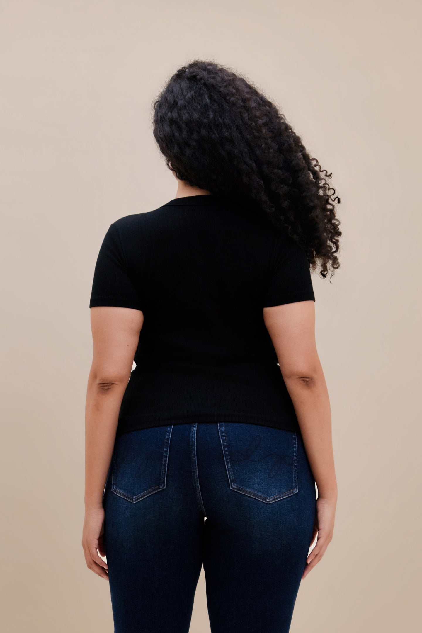 Backside of a woman wearing a fitted black rib tee and dark jeans against a beige wall 
