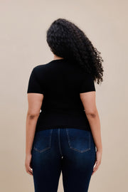 Backside of a woman wearing a fitted black rib tee and dark jeans against a beige wall 