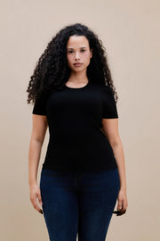 Woman wearing a fitted black rib tee and dark jeans against a beige wall 