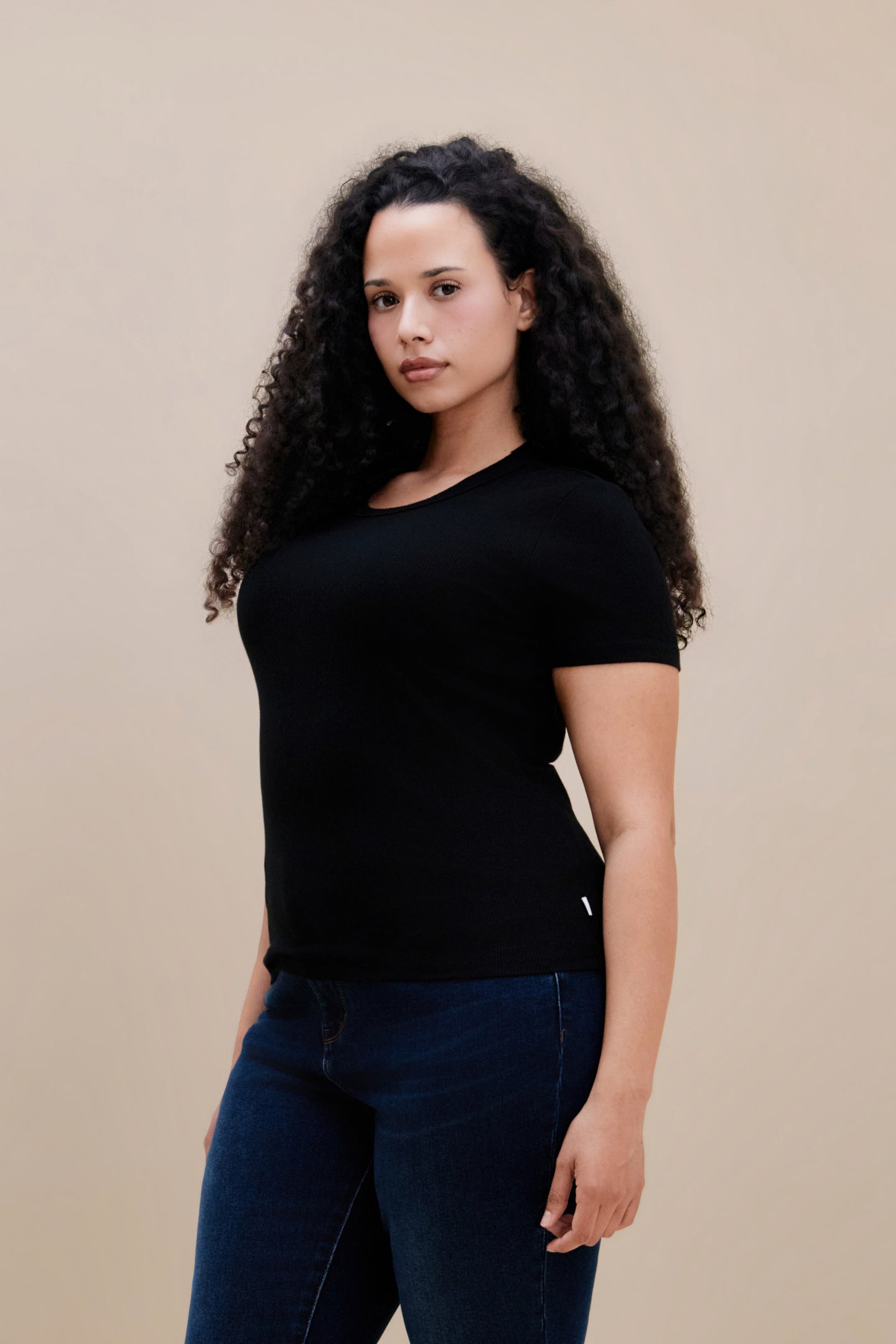 Woman wearing a fitted black rib tee and dark jeans against a beige wall 