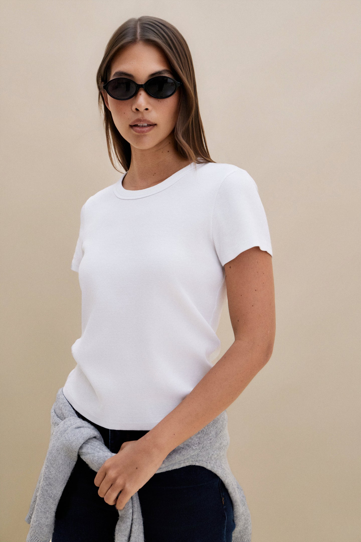 Woman wearing a white t-shirt, gray cardigan, and sunglasses against a beige background