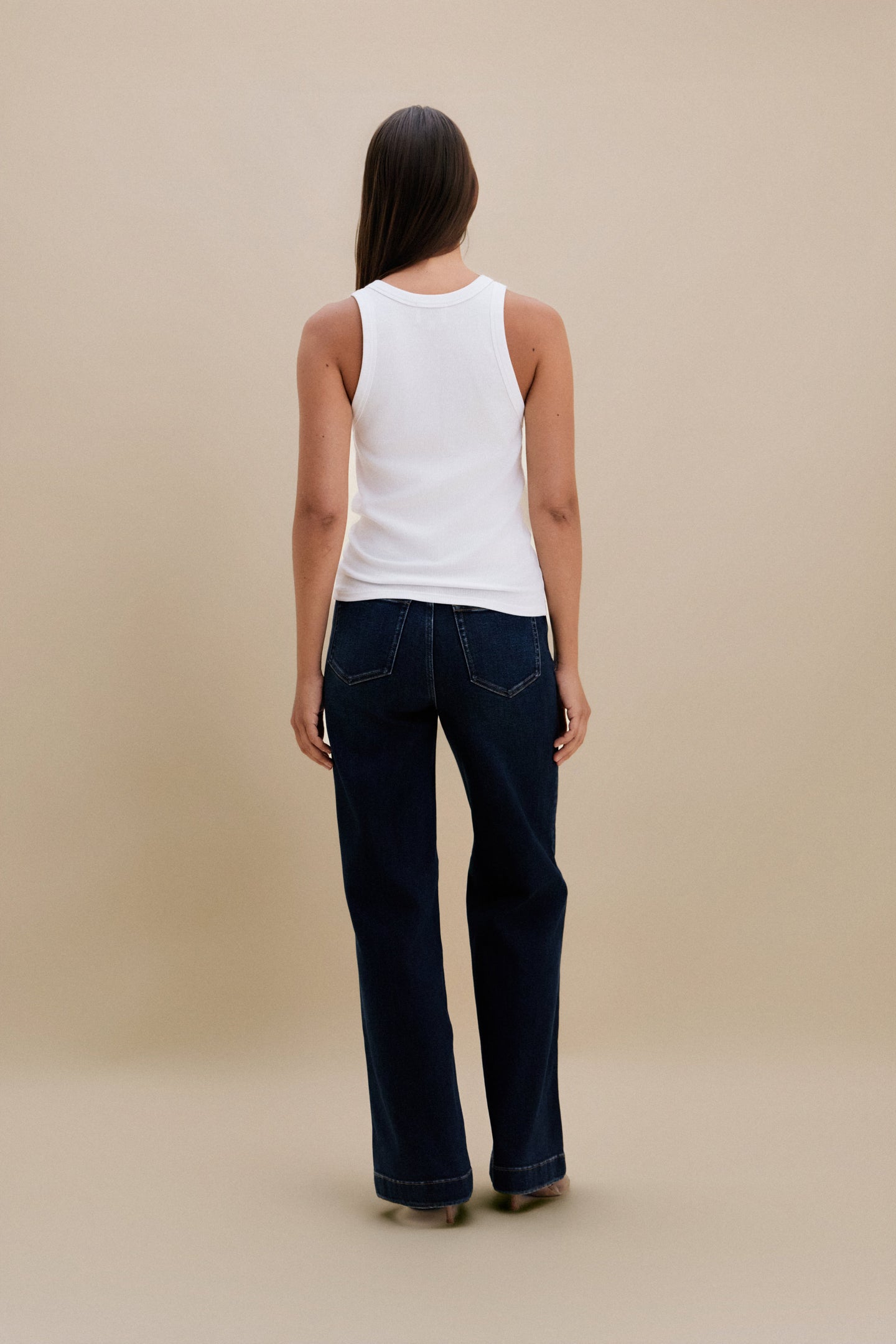 Person wearing a white tank top and dark blue jeans against a beige background