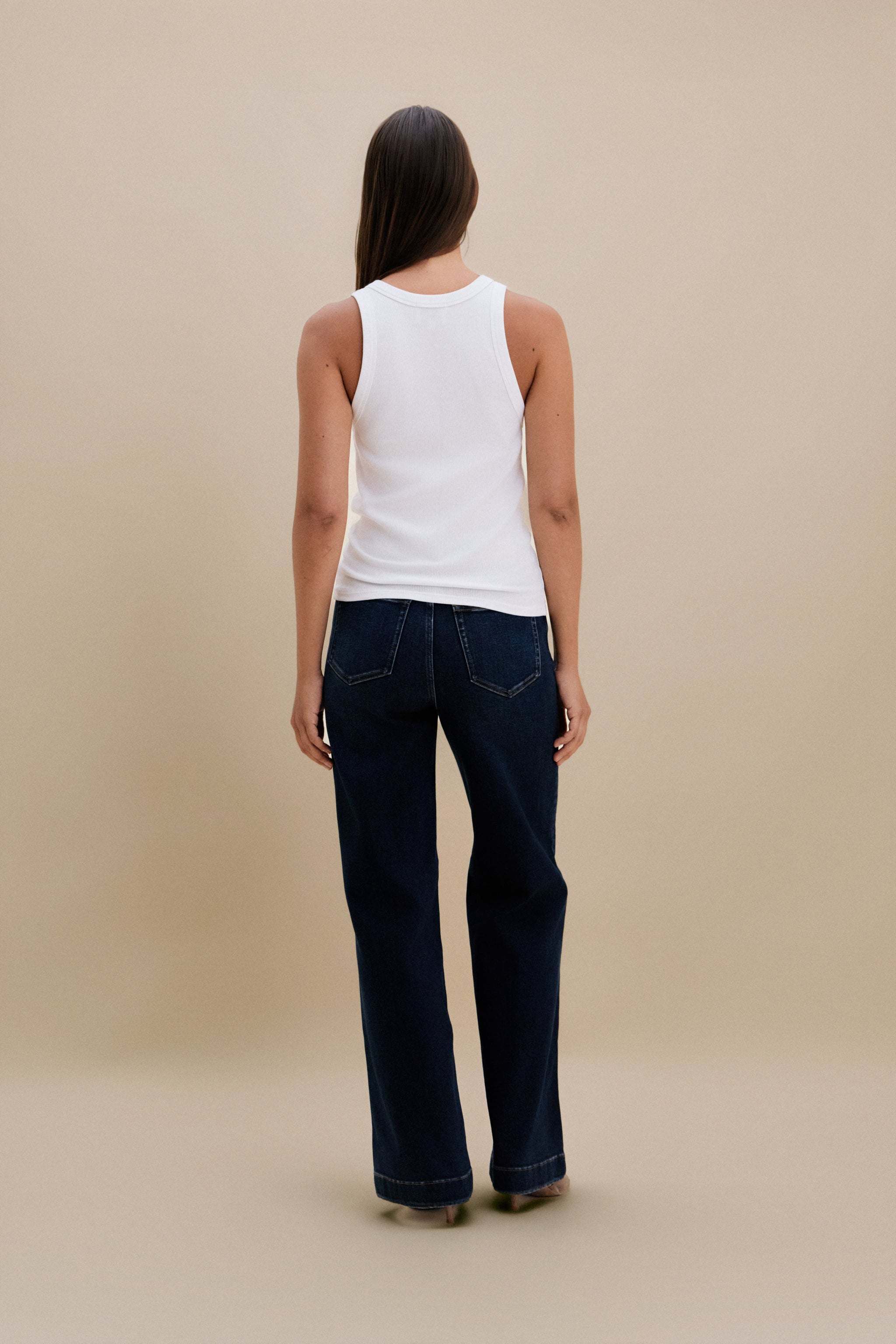 Person wearing a white tank top and dark blue jeans against a beige background