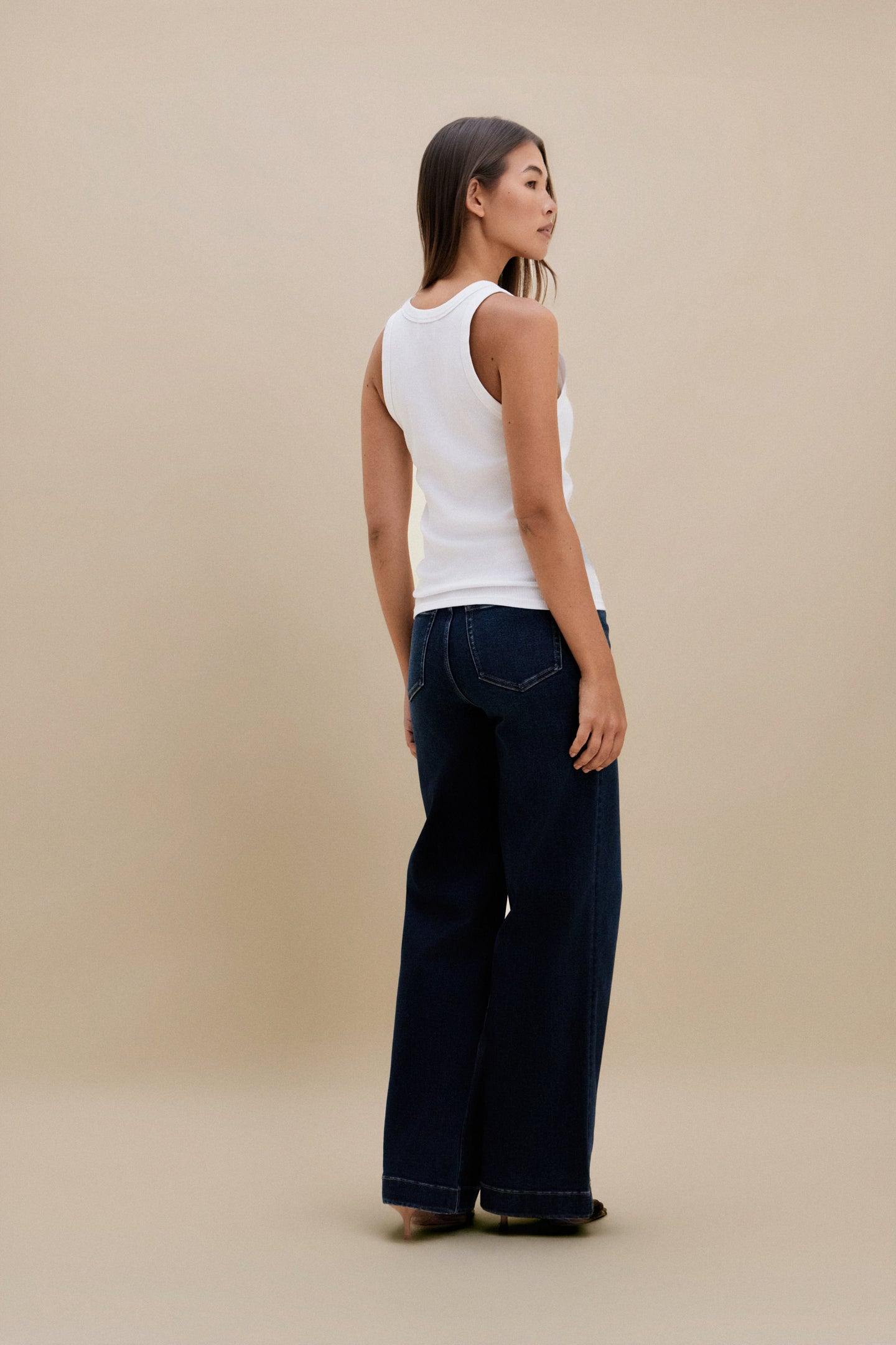 Woman wearing a white sleeveless top and dark blue jeans against a beige background