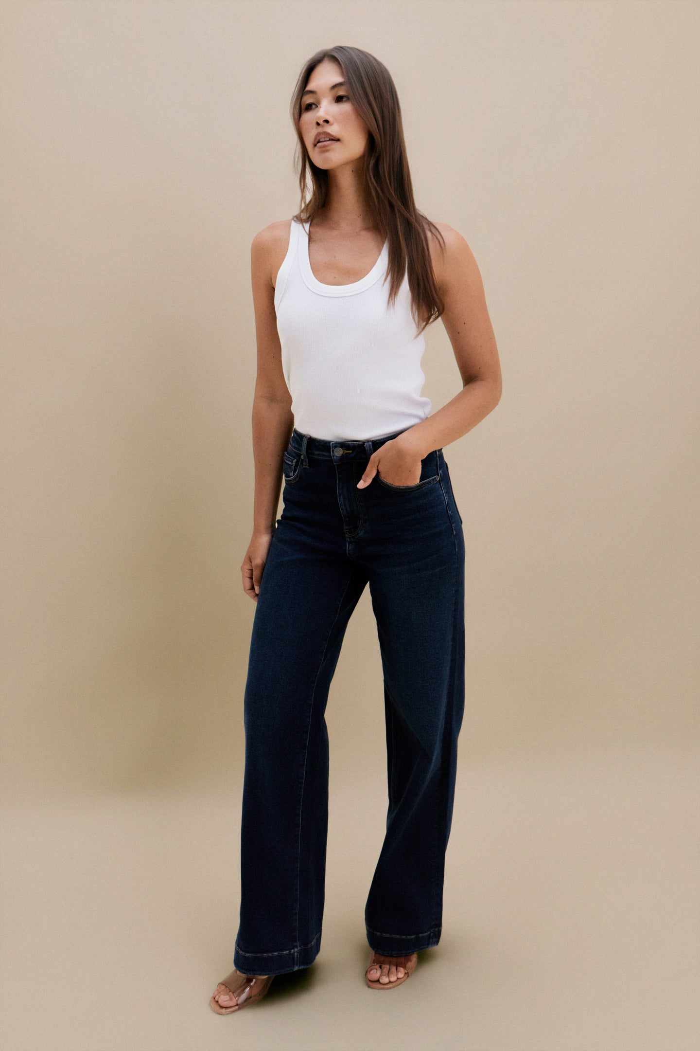 Woman wearing a white tank top and dark blue jeans against a beige background