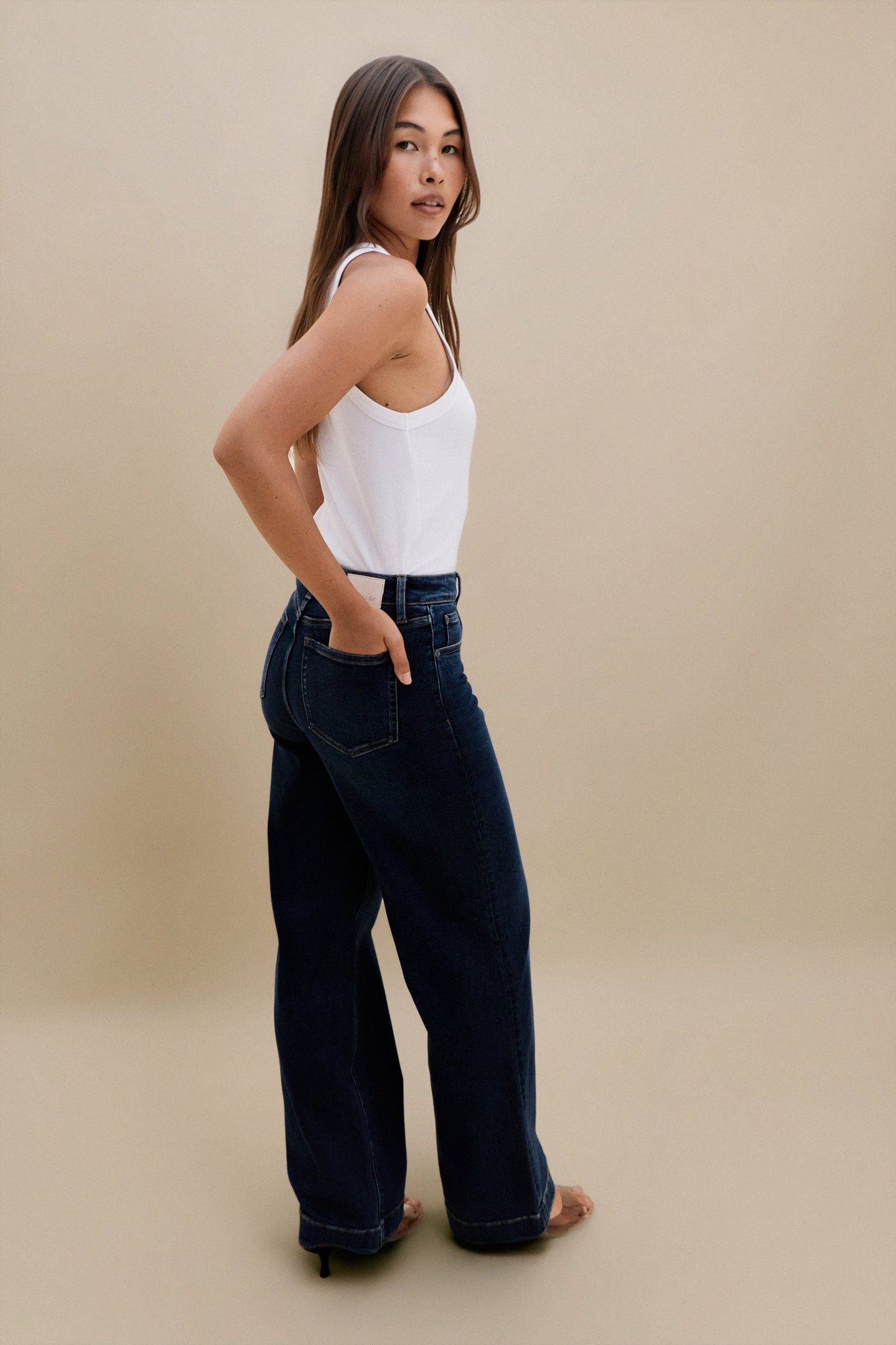 Woman wearing a white tank top and dark blue jeans against a beige background