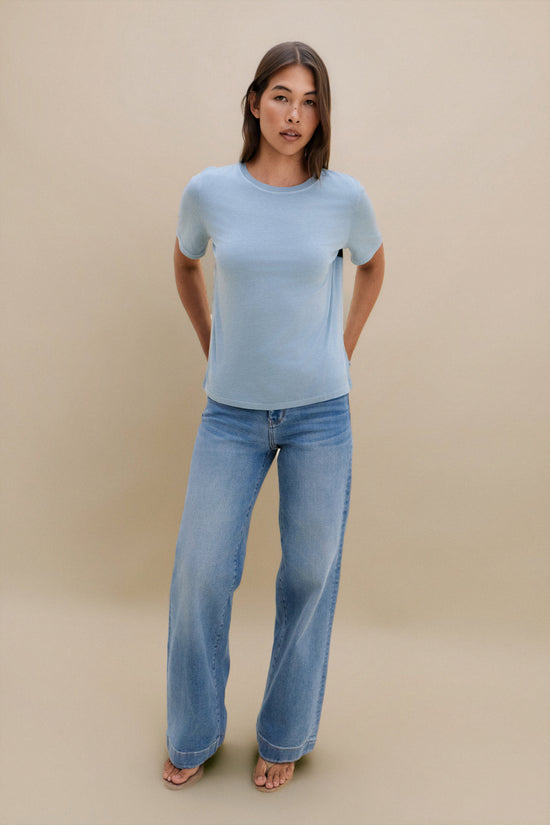 Woman wearing a light blue top and jeans against a beige background 