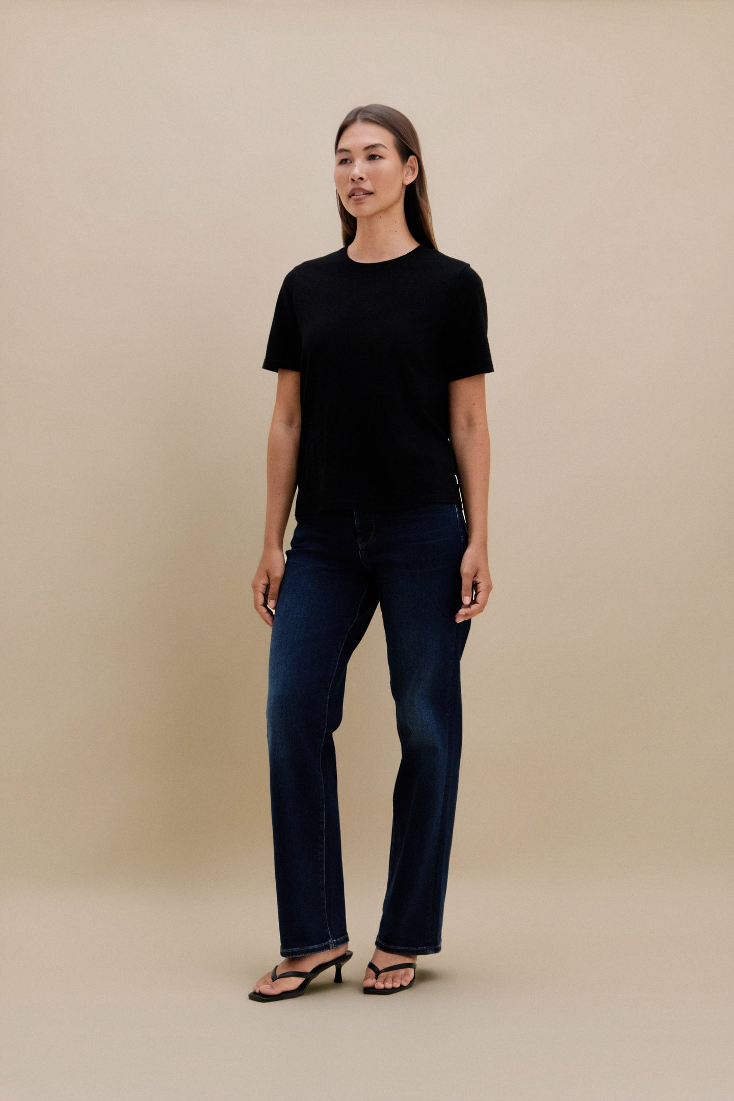 Woman wearing a black crew neck t-shirt and blue jeans standing in front of a beige wall 