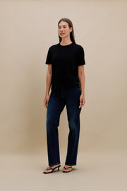 Woman wearing a black crew neck t-shirt and blue jeans standing in front of a beige wall 