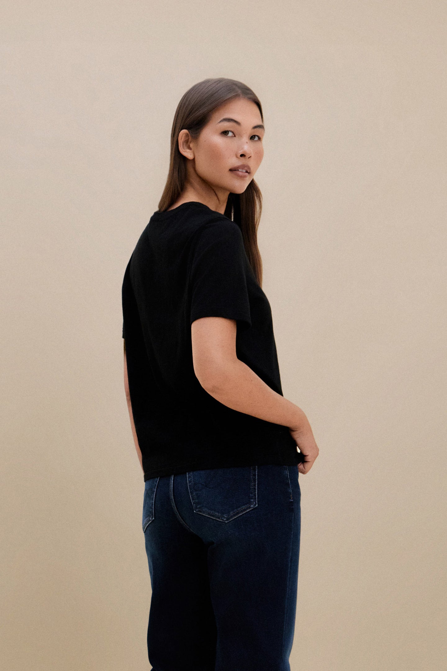 Woman wearing a black crew neck t-shirt and blue jeans standing in front of a beige wall 