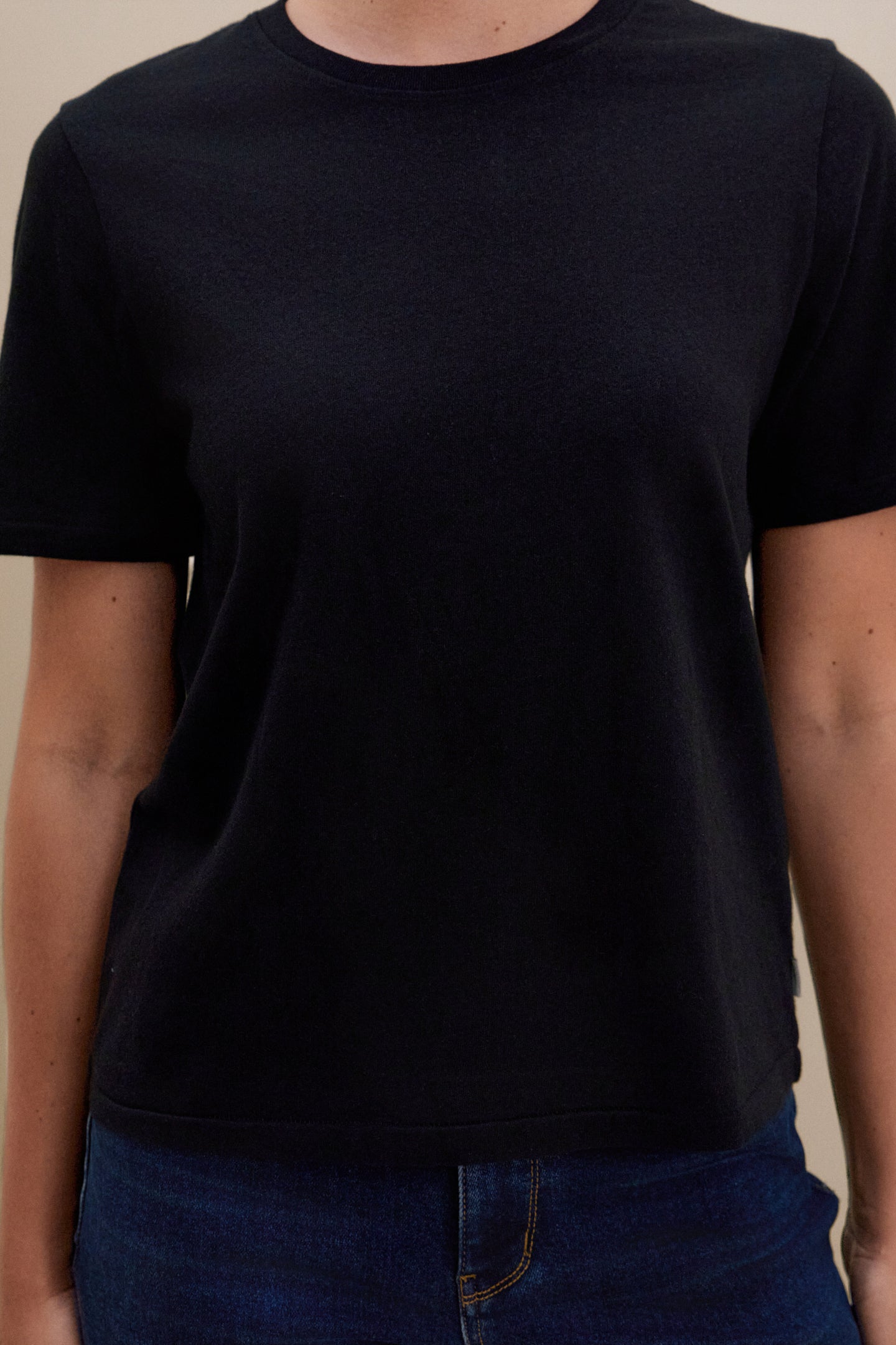 Woman wearing a black crew neck t-shirt and blue jeans standing in front of a beige wall 