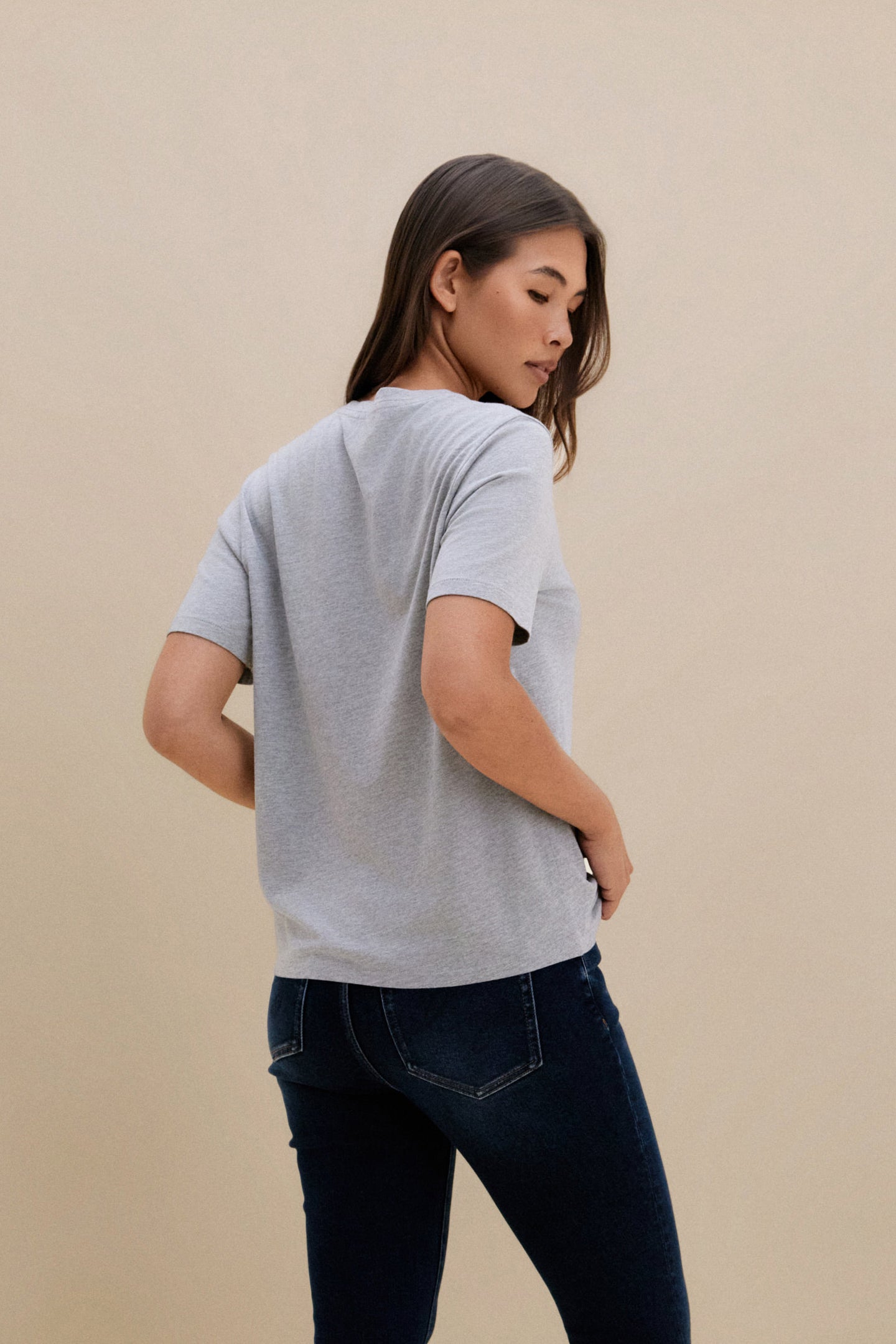 Woman wearing a gray t-shirt and dark jeans against a beige background