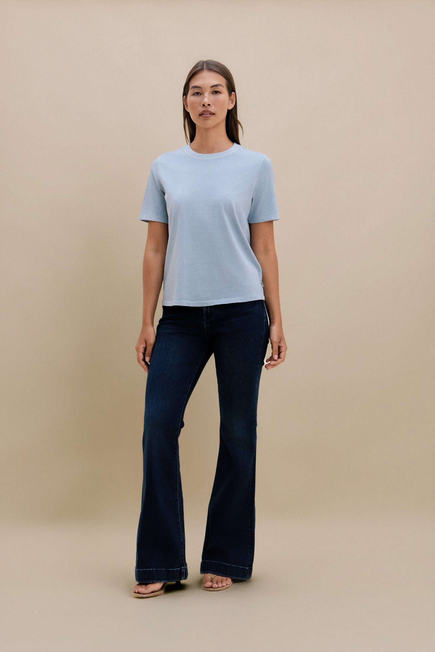 Woman wearing a light blue t-shirt and dark jeans against a beige background