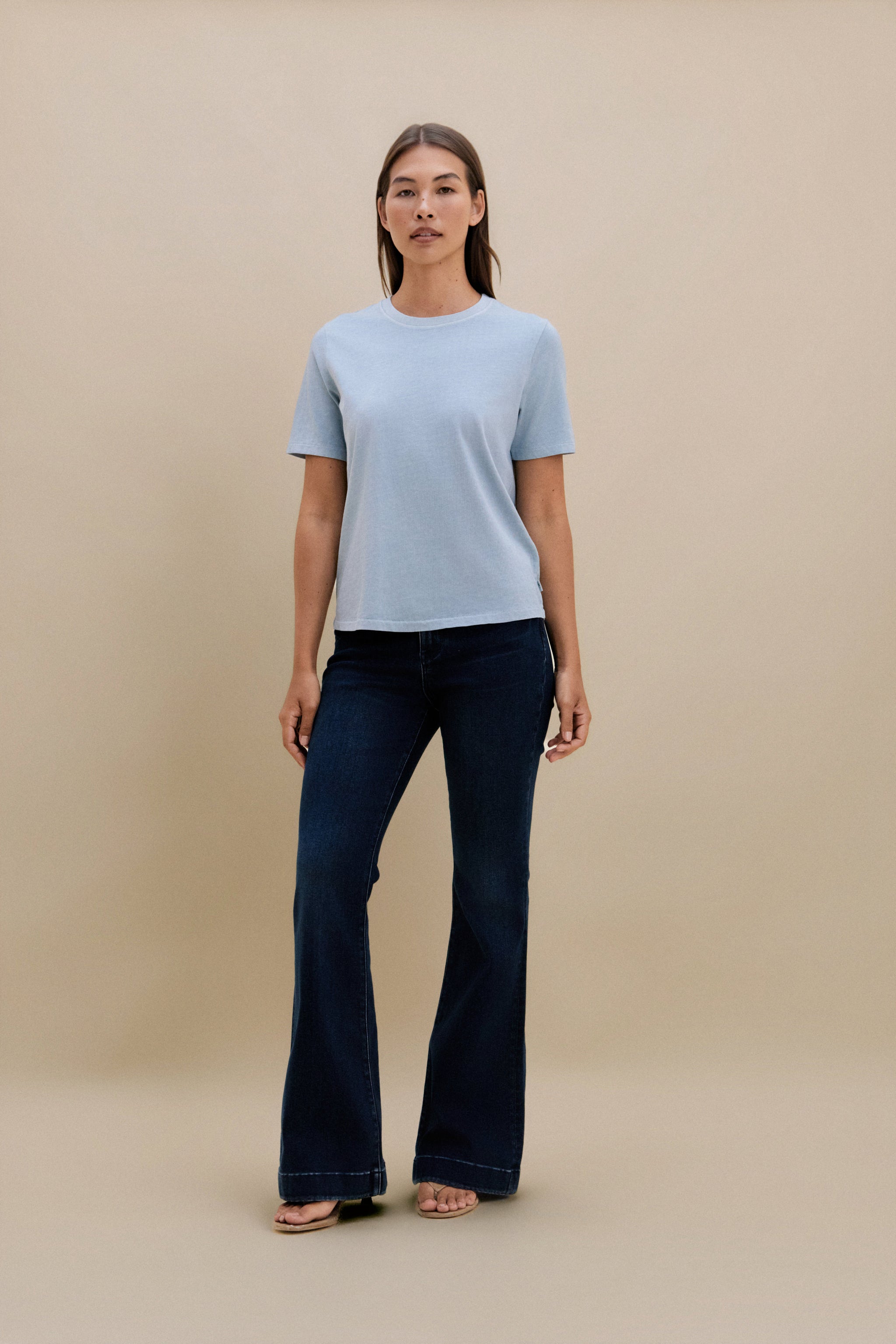 Woman wearing a light blue t-shirt and dark jeans against a beige background