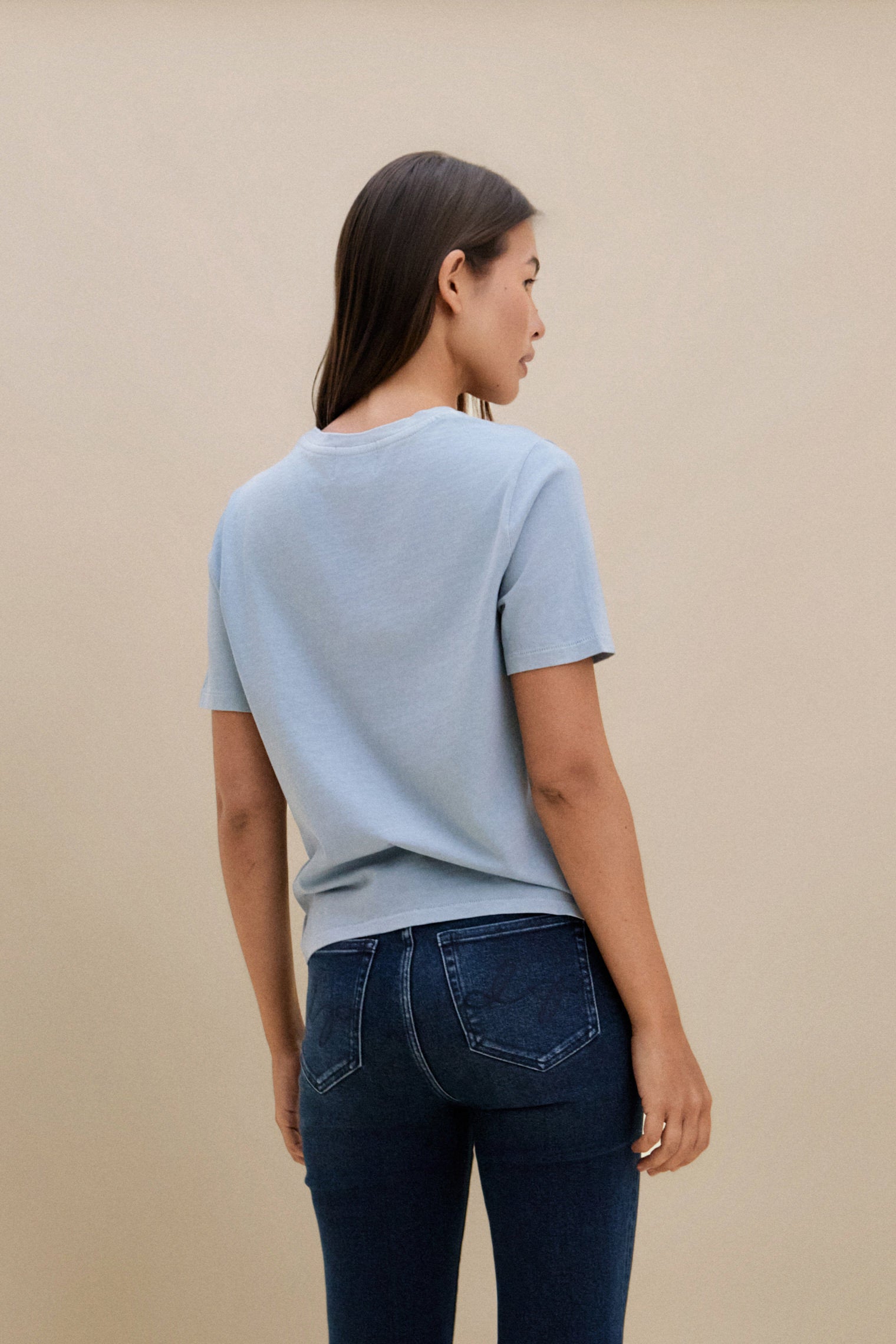 Woman wearing a light blue t-shirt and dark jeans against a beige background