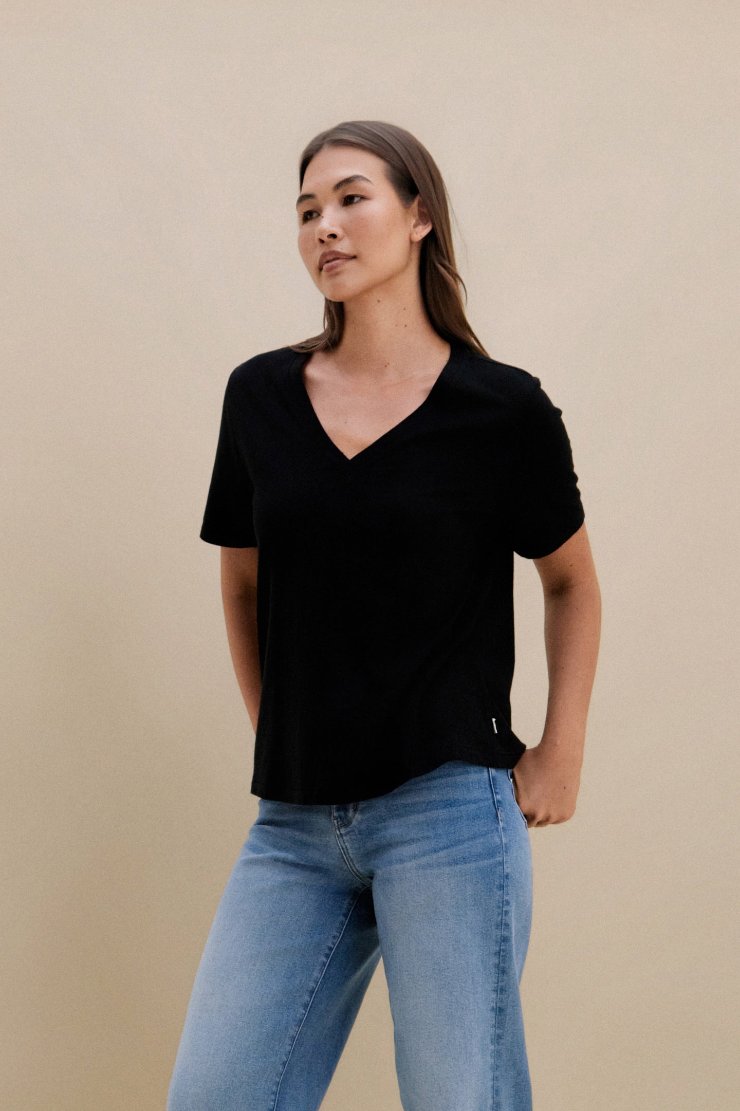 Woman wearing a black v-neck t-shirt and blue jeans against a beige wall 