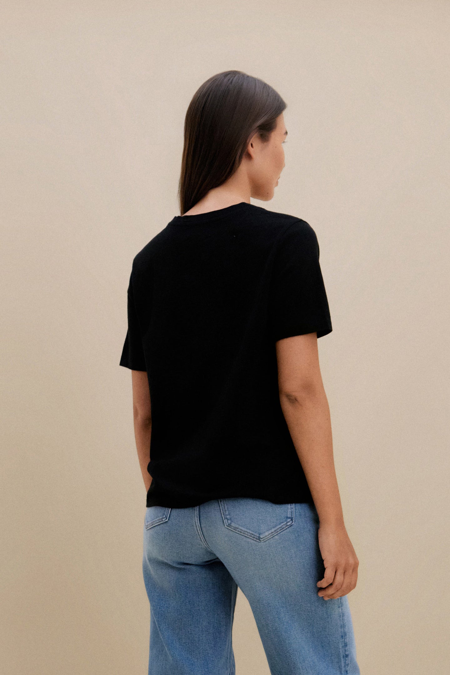 Woman wearing a black v-neck t-shirt and blue jeans against a beige wall 