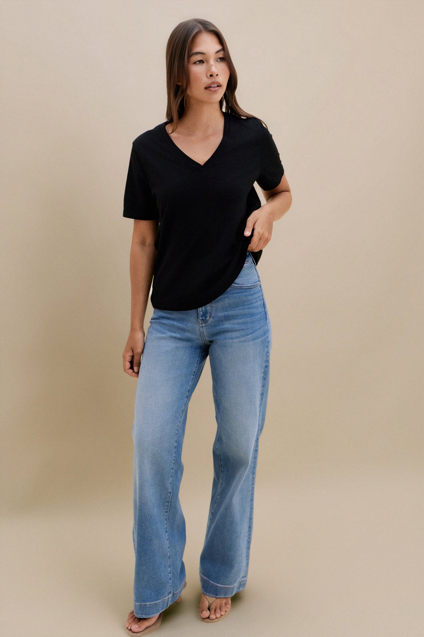 Woman wearing a black v-neck t-shirt and blue jeans against a beige wall 