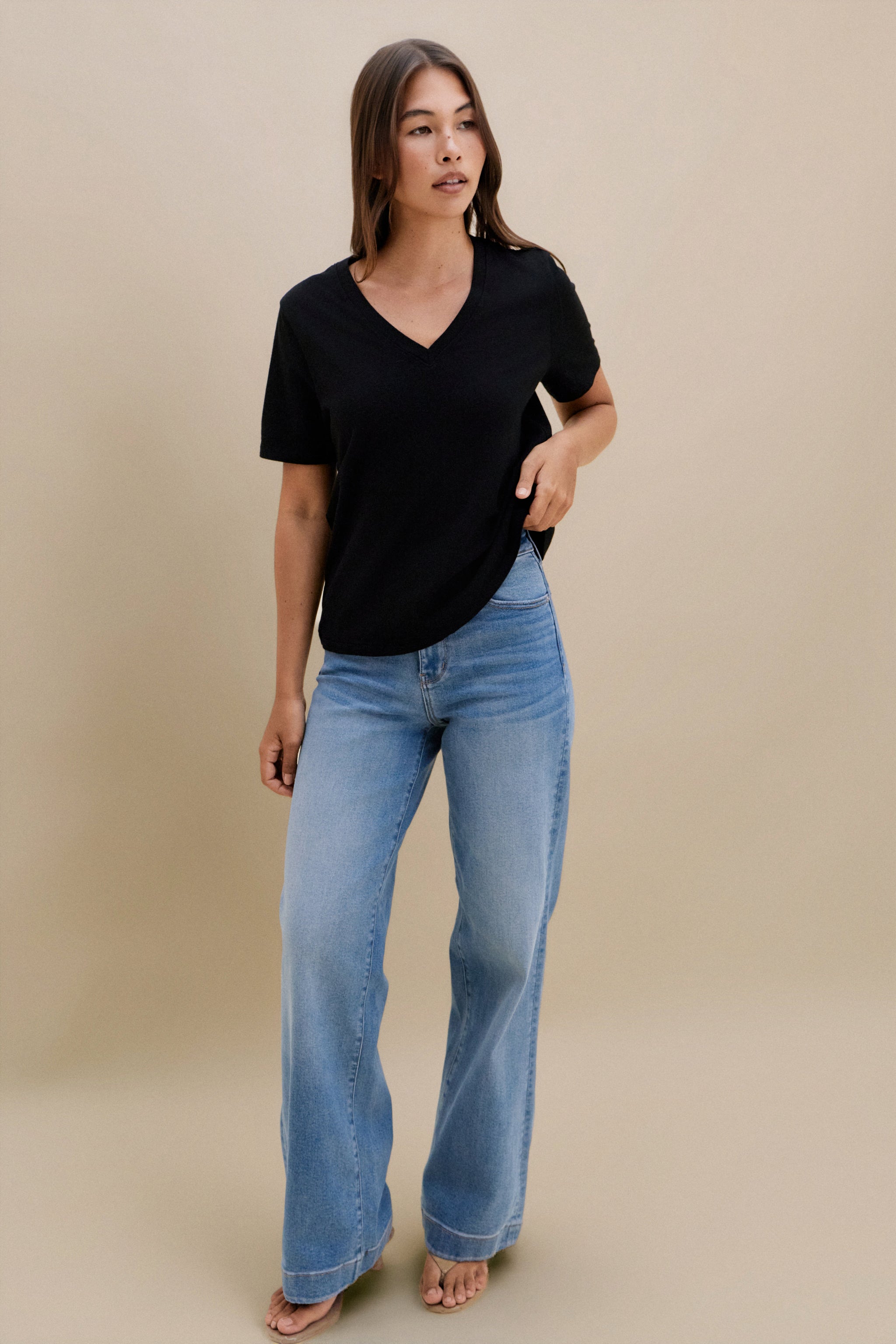 Woman wearing a black v-neck t-shirt and blue jeans against a beige wall 