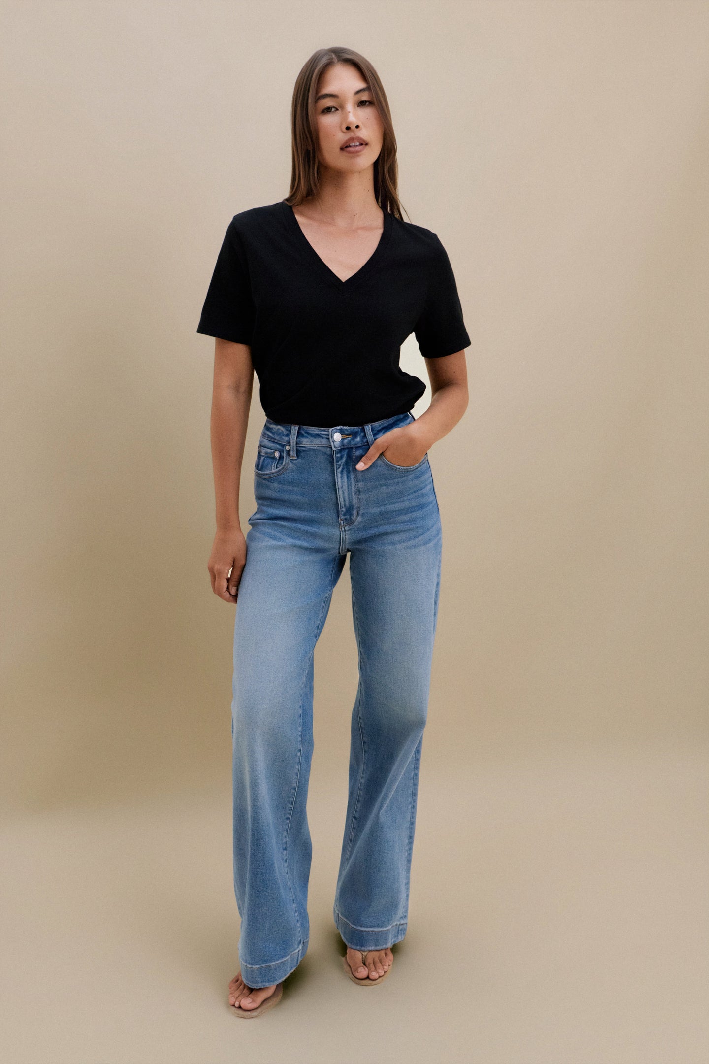 Woman wearing a black v-neck t-shirt and blue jeans against a beige wall 