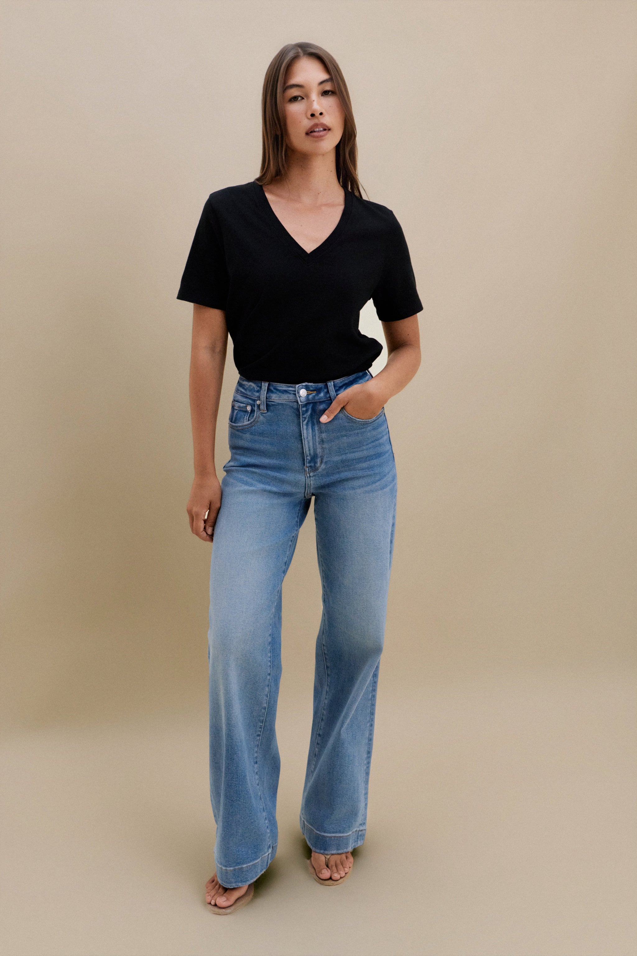 Woman wearing a black v-neck t-shirt and blue jeans against a beige wall 