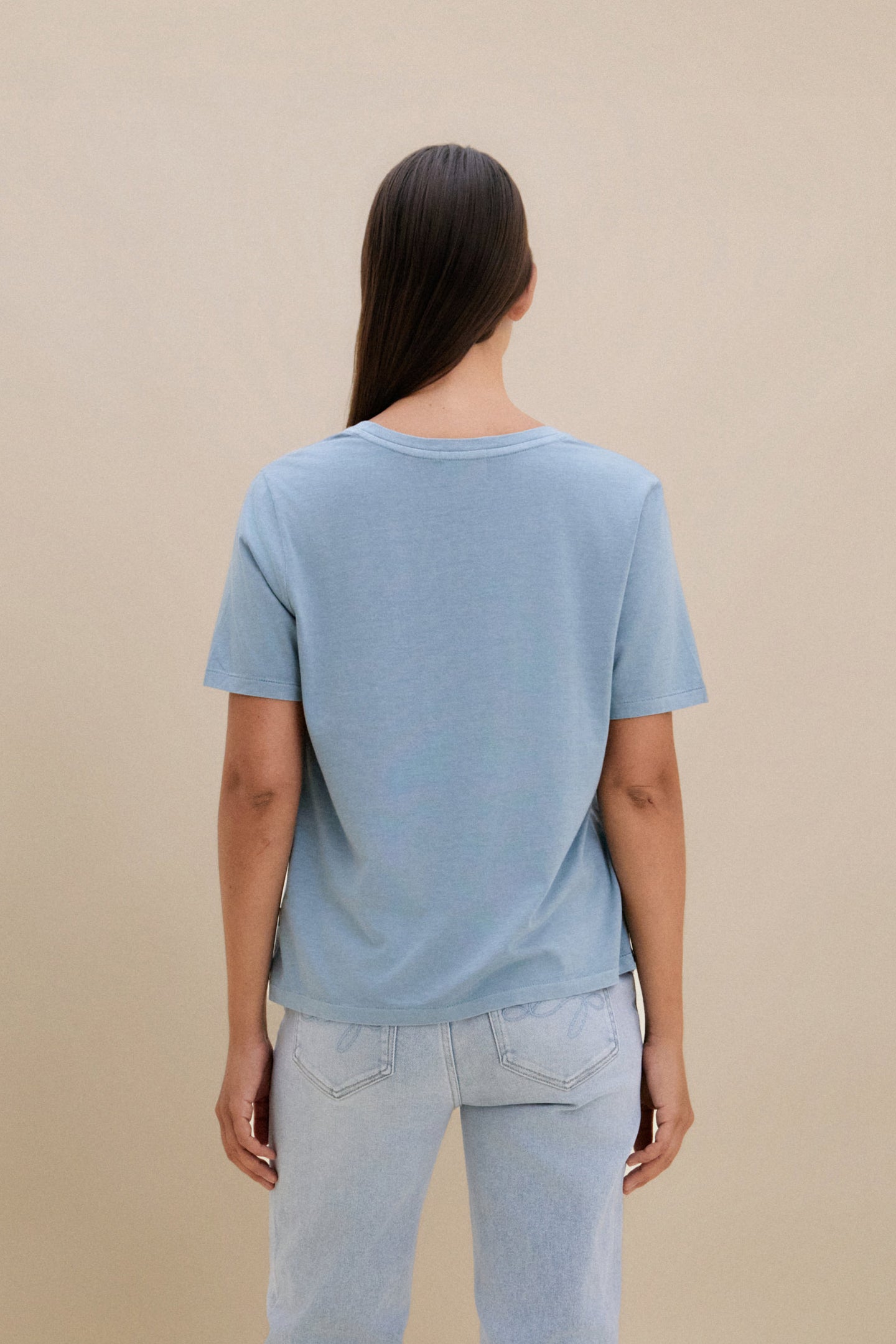 woman wearing light blue t-shirt and light colored denim with a beige wall 